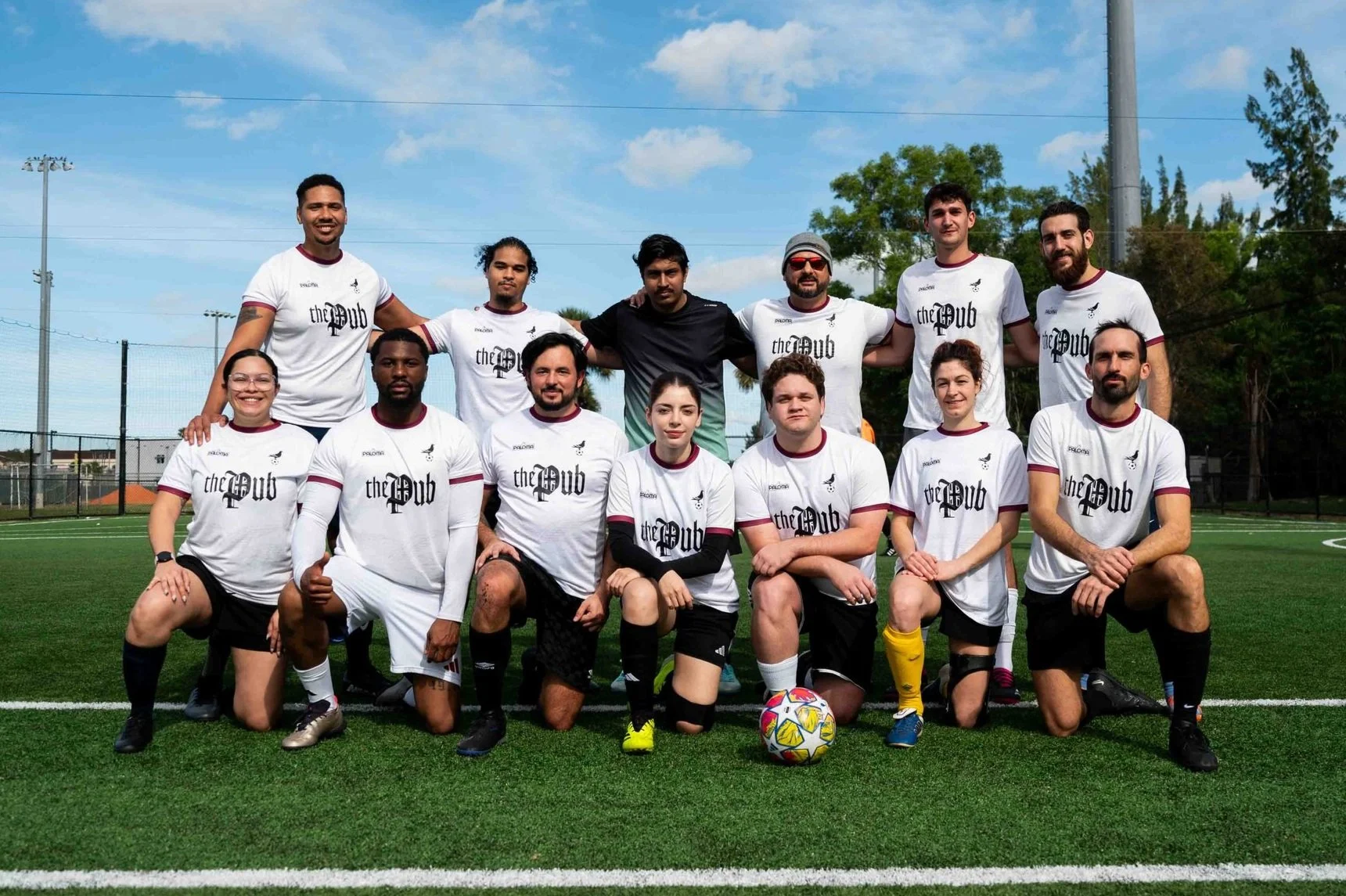 A group of 12 people on a soccer field posing for a team photo, with 6 people kneeling in front and 6 standing behind them, all wearing matching white soccer jerseys with