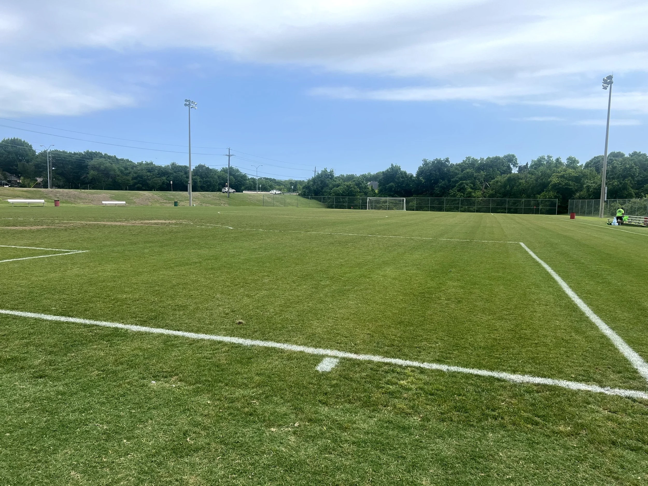 A view of a football field with green grass and white boundary lines, surrounded by sports lighting towers, trees, and a few cars on a distant road under a partly cloudy sky.