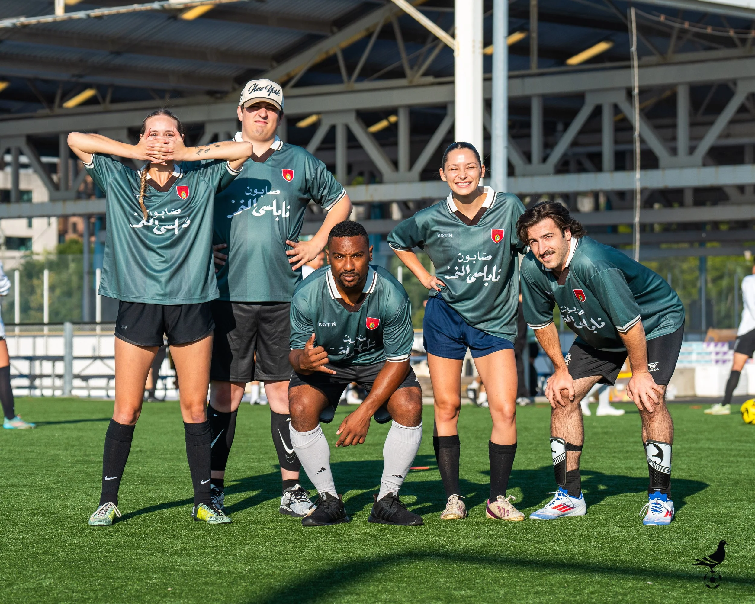 Group of five diverse soccer players posing on a green field, dressed in teal jerseys with white and black accents, some crouching and others standing, under a metallic structure, with a bright sunny sky in the background.