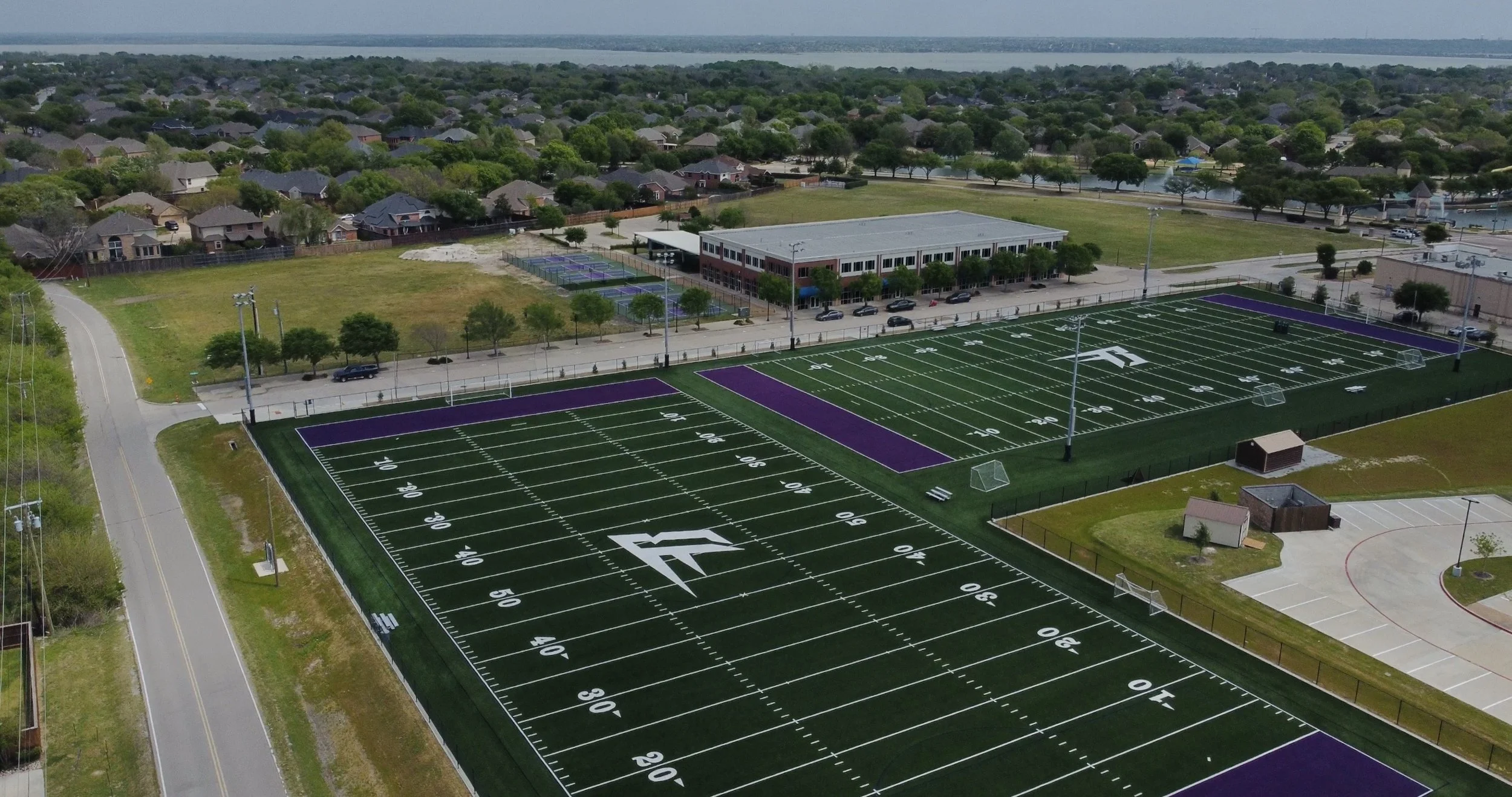 Aerial view of a football field with purple end zones, surrounded by a parking lot, adjacent to a school building, with tennis courts nearby. Residential houses and a body of water are visible in the background.