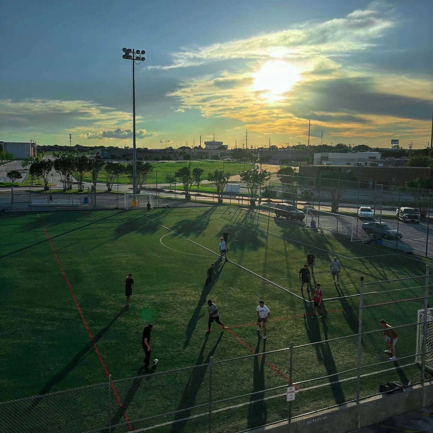 People playing soccer on a field during sunset, with trees, a parking lot, and a cloudy sky in the background.