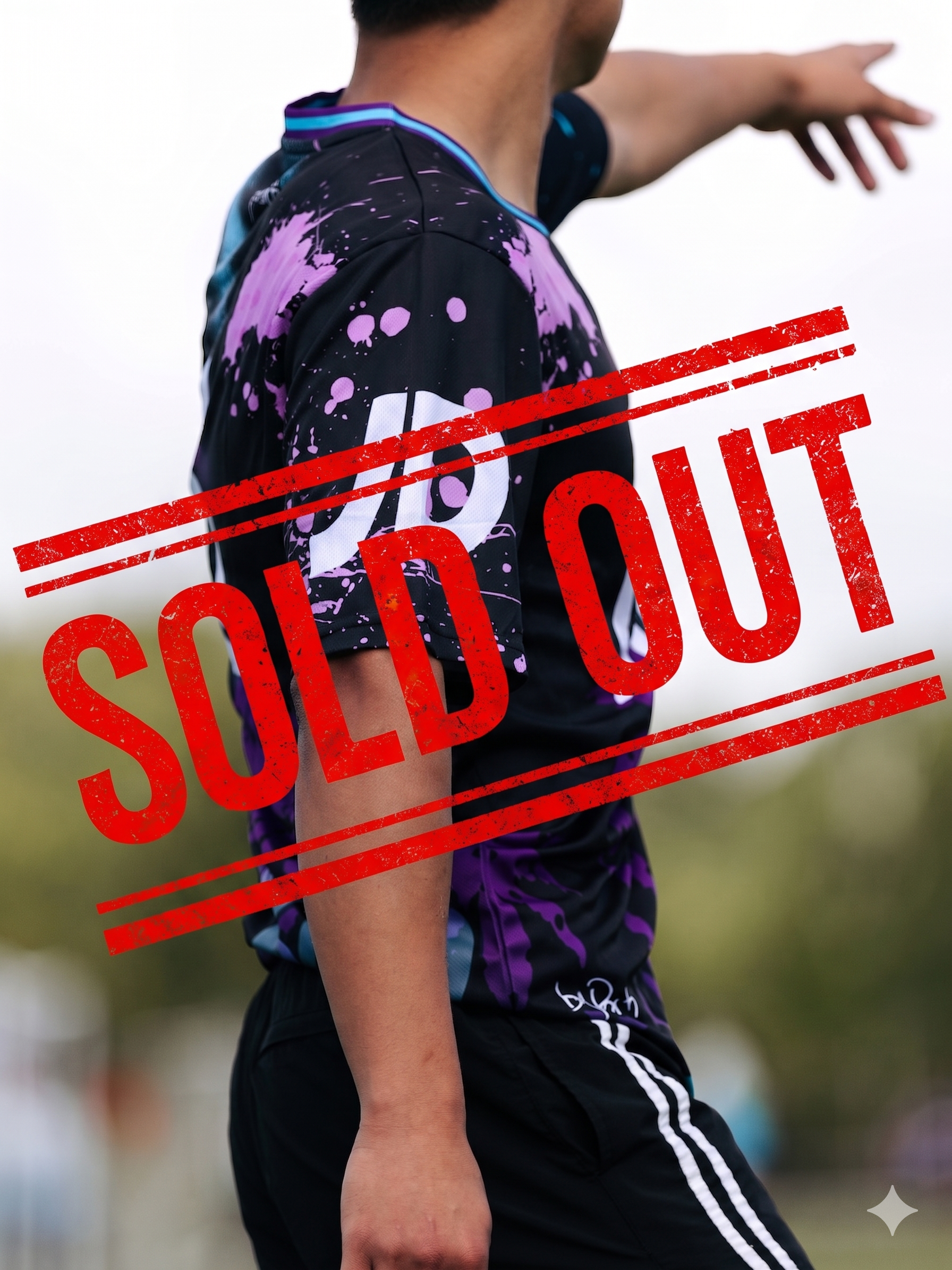 Close-up of a person wearing a black and purple sports jersey with white and purple design, and black shorts with white stripes, extending arm outward with fingers pointing, seen outdoors with a blurred green field background. Red 'SOLD OUT' stamp overlays the image.