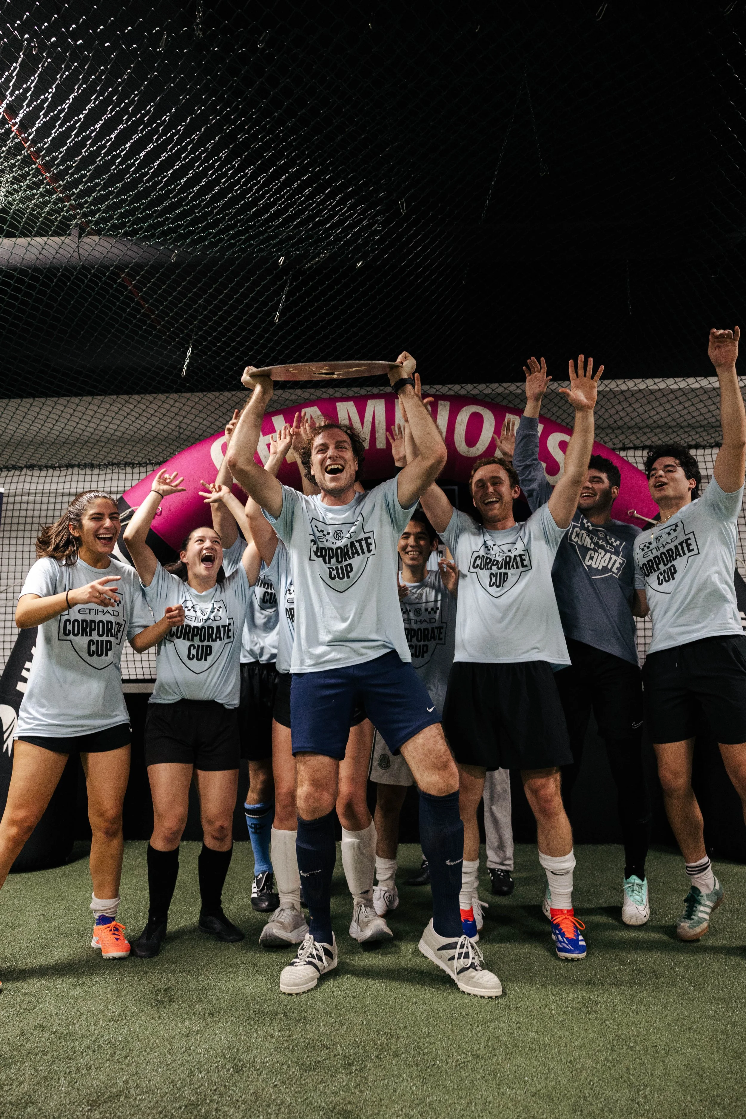 A group of people celebrating a sports victory indoors, with a man in the center holding a trophy above his head. They are wearing sports uniforms with a