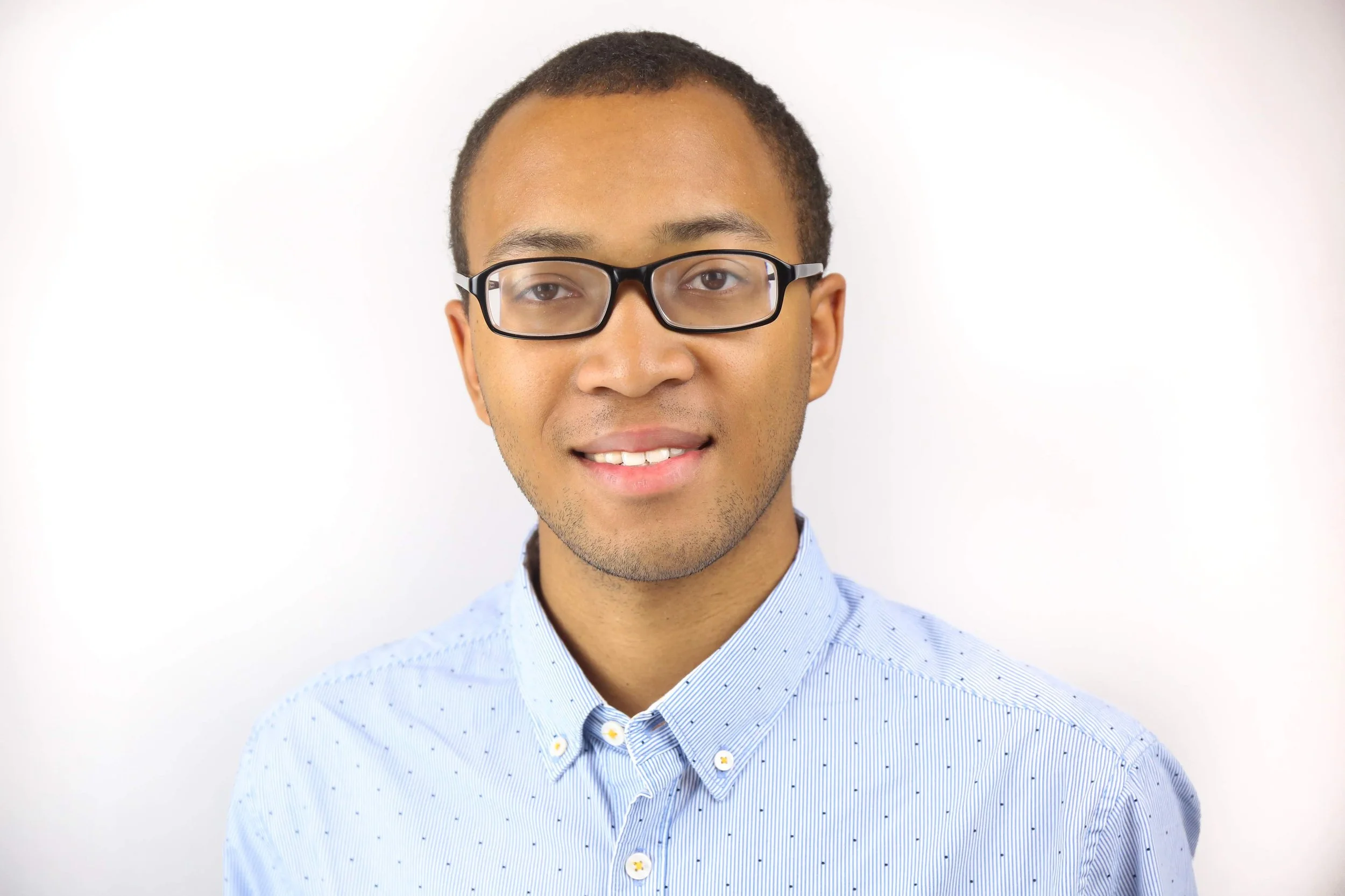 Portrait of a young man with glasses, wearing a light blue button-up shirt, smiling against a plain white background.