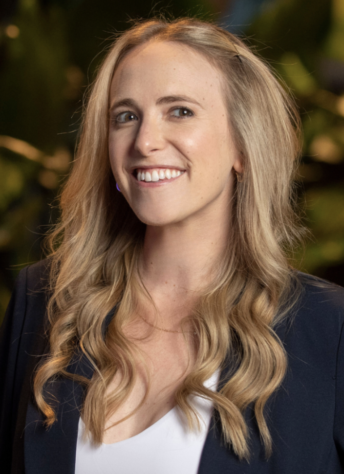 A woman with long blonde wavy hair smiling, wearing a navy blazer over a white top, with a blurred green background.