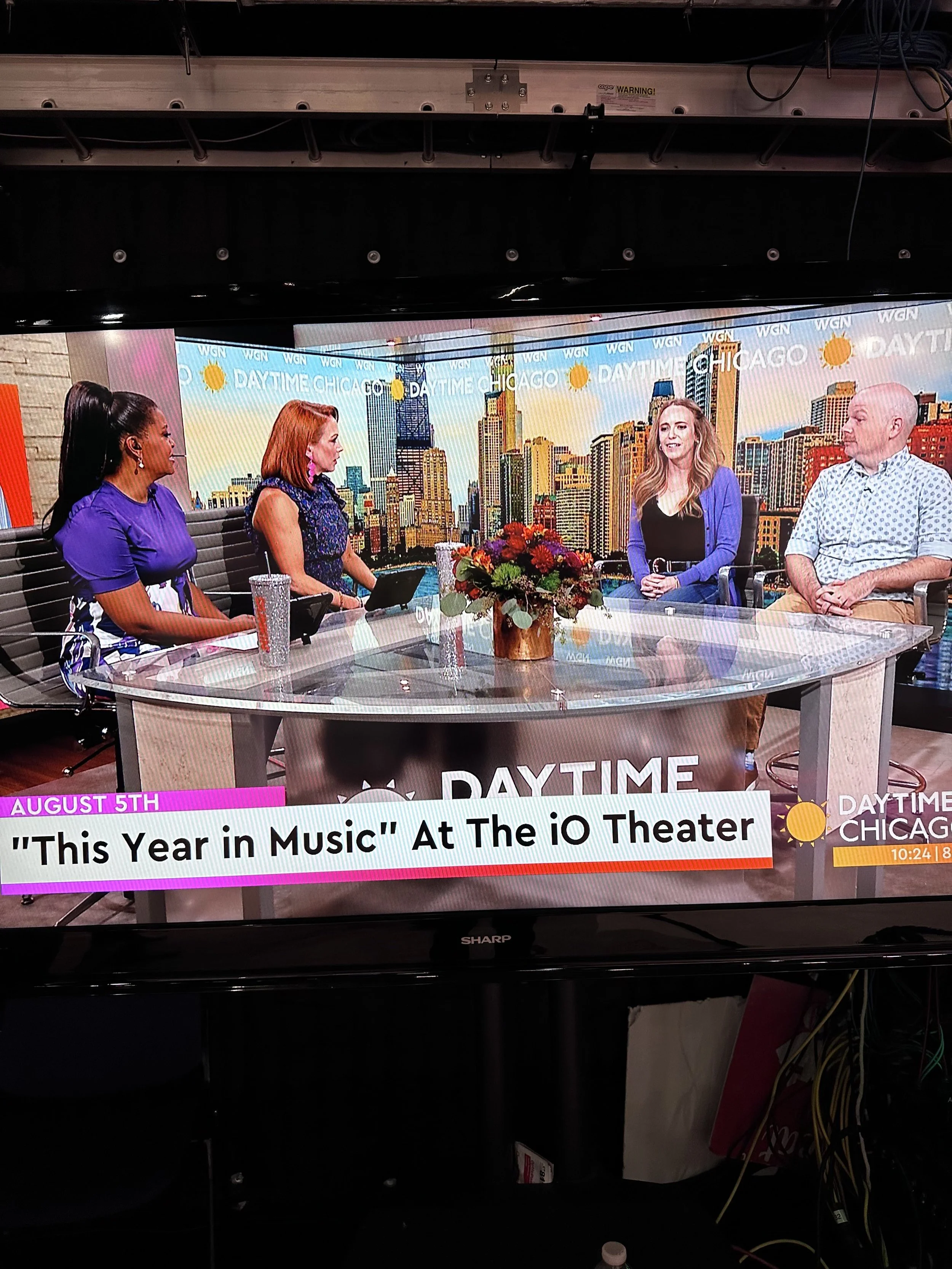 Television screen showing a live broadcast of a daytime talk show with four women sitting around a table with a cityscape background. The show is titled 'DAYTIME CHICAGO' and the topic is 'This Year in Music' at The IO Theater, with the date August 5th displayed.