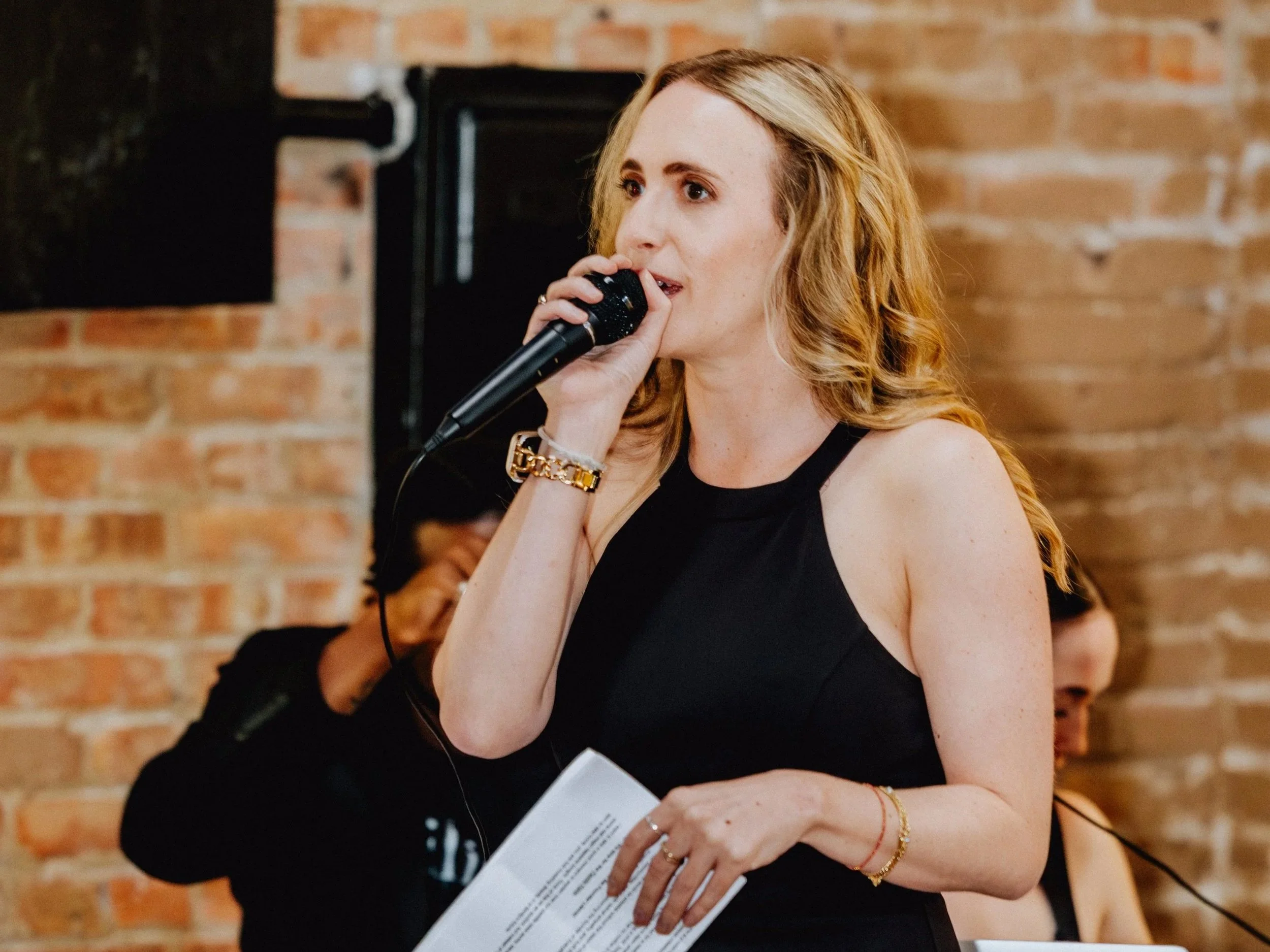 A woman with red hair holding a microphone and a sheet of paper, standing in front of a brick wall. She appears to be speaking or singing.
