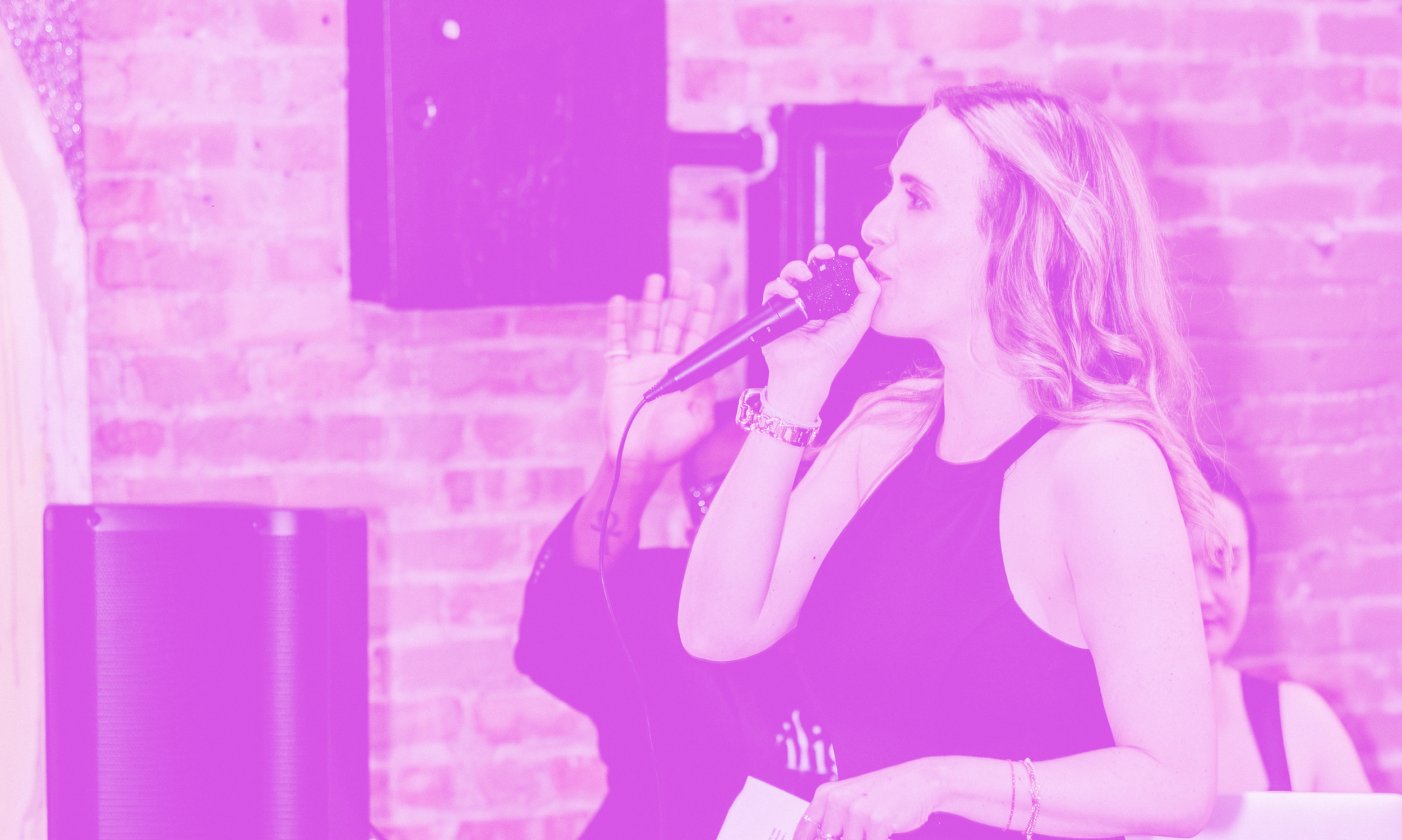 Woman speaking into a microphone in a room with a brick wall background, hosting a non-profit gala