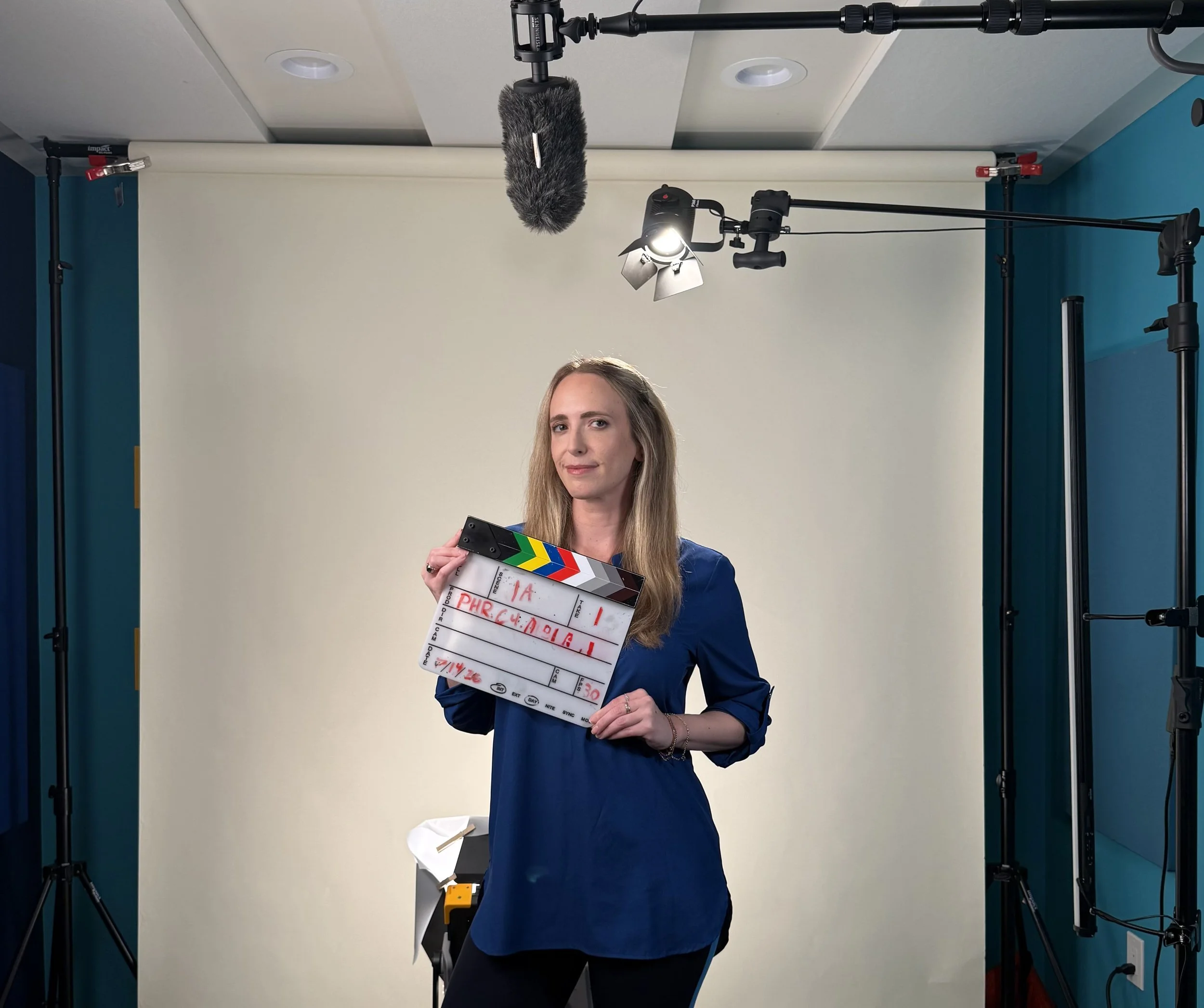 A woman with long blonde hair standing in front of a filming backdrop, holding a clapperboard with red writing, surrounded by professional filming equipment including lights, a boom microphone, and stand supports.