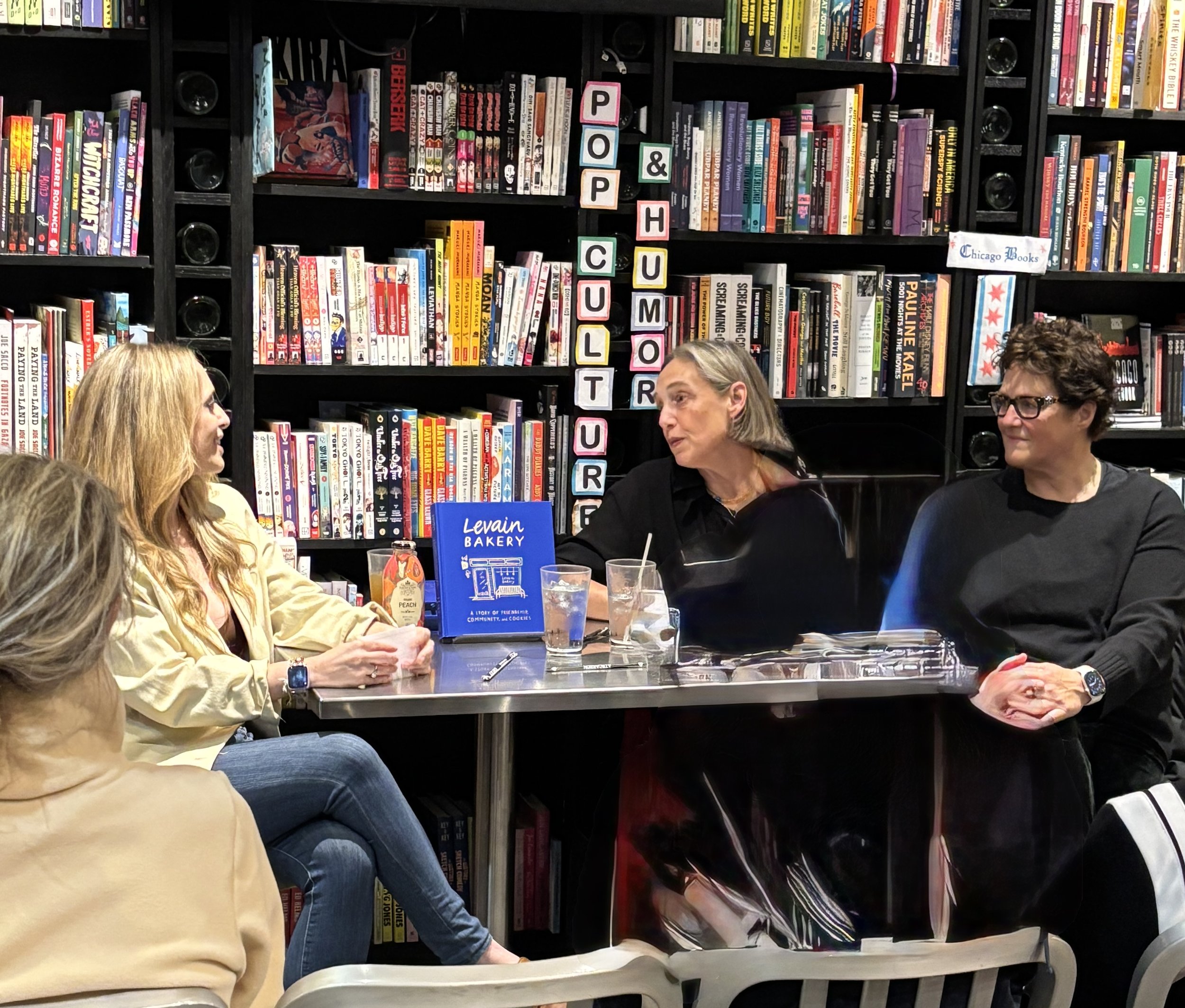 A gathering of people in a bookstore or library with bookshelves in the background. There are three women in the foreground, one with blonde hair wearing glasses, another with gray hair, and a third with dark hair and glasses. Items like glasses, drinks, and a blue sign called "Levain Bakery" are on the table. The background shows colorful books and signs with letters spelling out "POP & CULTURE".
