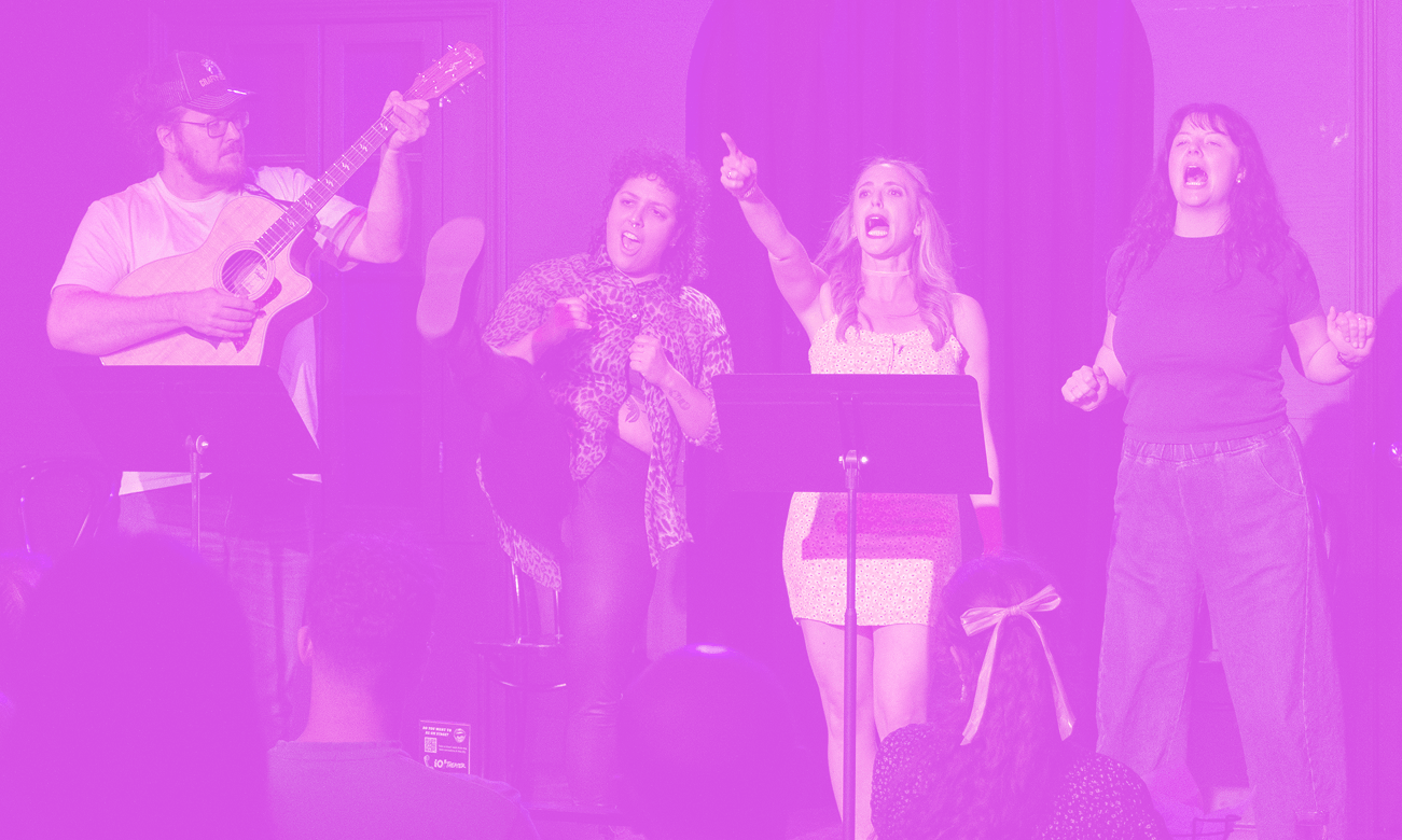 A group of four people performing on stage: a man playing guitar, and three women singing passionately, one of whom is pointing upward.