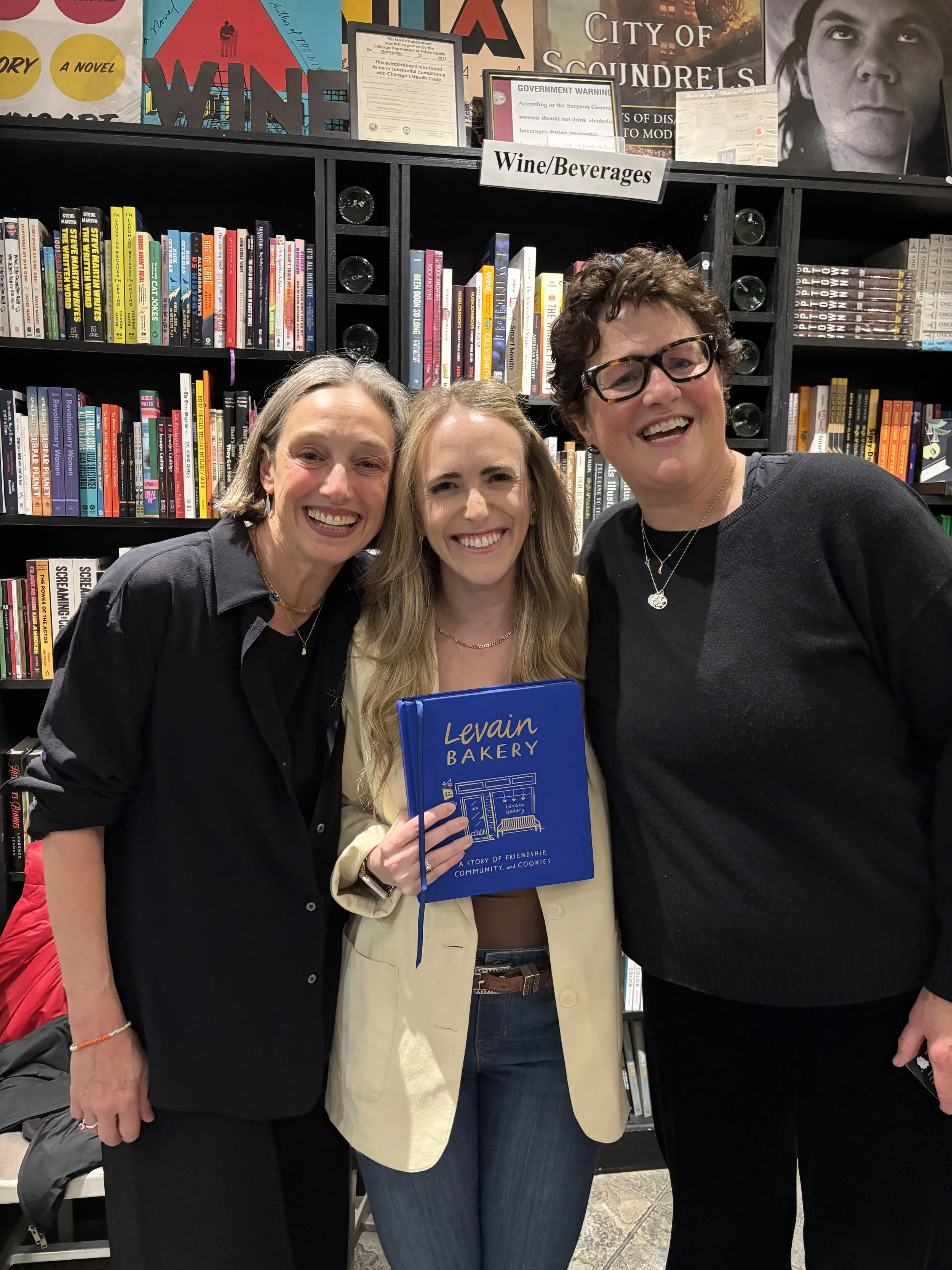 Three women standing in front of a bookshelf filled with books, smiling and posing for the photo. One woman in the middle is holding a blue book titled 'Levin Bakery'.