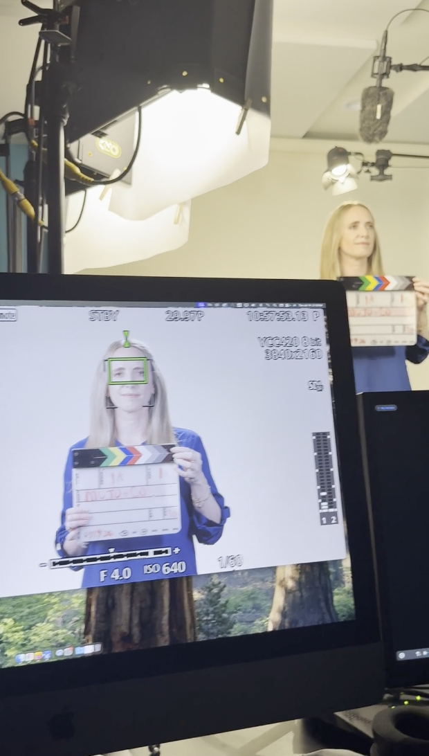 A woman is standing in front of a gray backdrop holding a clapperboard, with her face and upper body visible, during a video shoot or recording studio. The scene is being captured on a monitor.