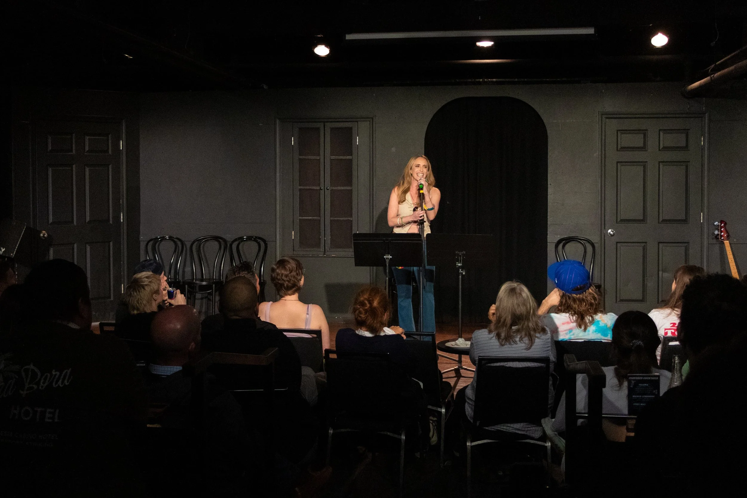 A woman on stage singing into a microphone during a performance or comedy show with an audience watching.