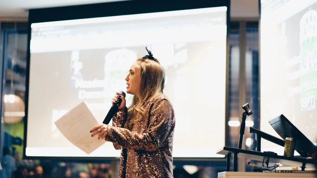 A woman speaking into a microphone while holding papers, standing in front of a large screen during a presentation or event.
