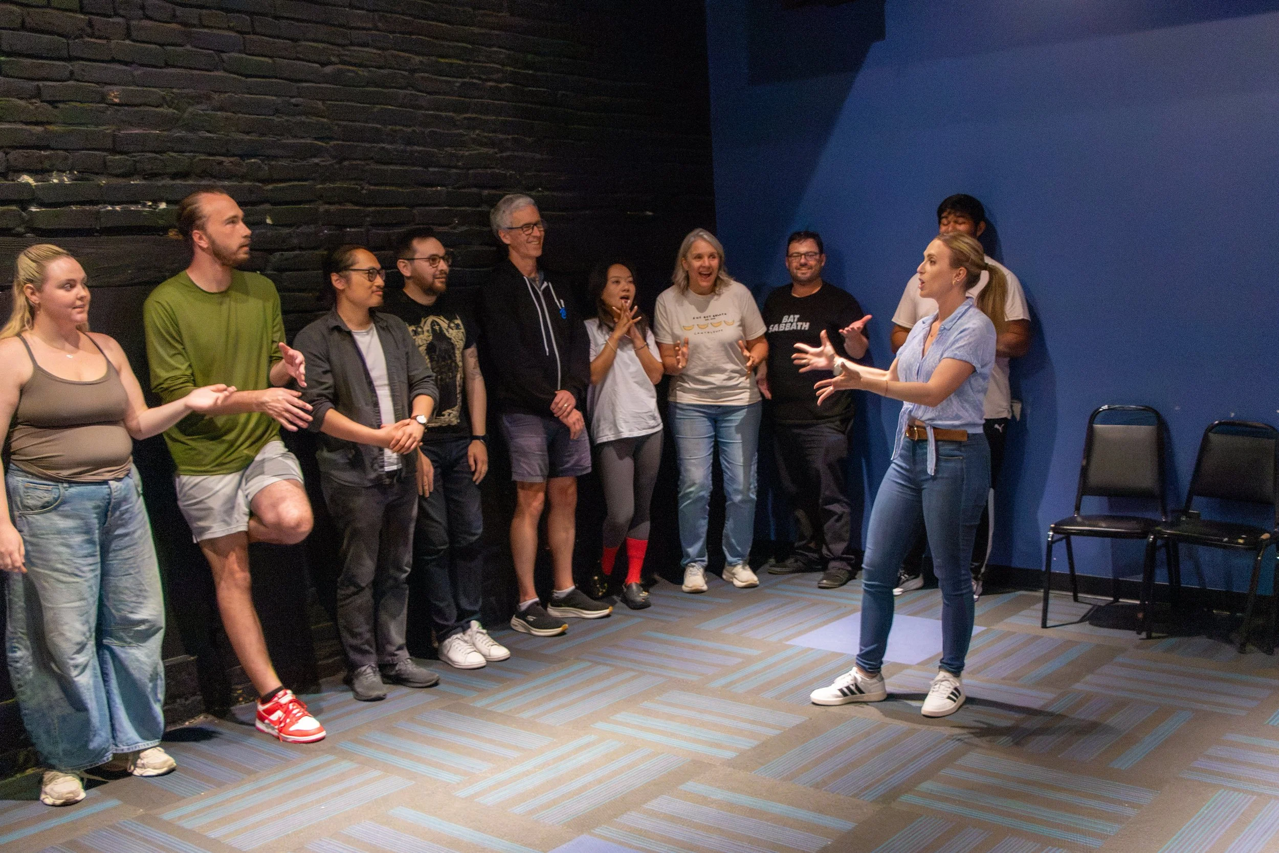 A woman in a light blue shirt and jeans is speaking or giving instructions to a group of approximately ten diverse people standing against a dark and blue wall. The group appears engaged and happy, with some smiling and clapping.