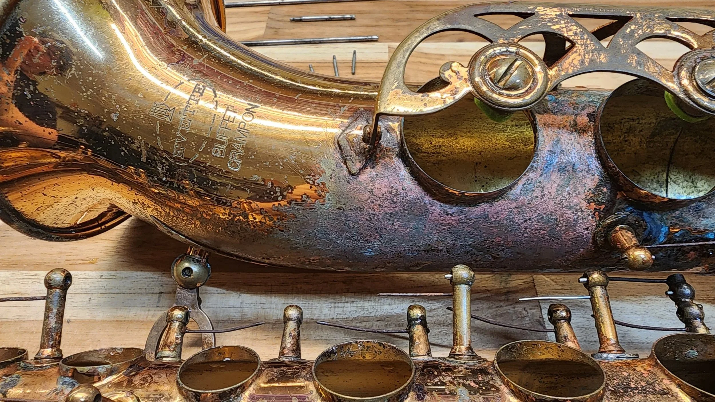 Close-up of a vintage gold-colored saxophone with visible scratches and tarnish, placed on a wooden surface with metal parts and screws around it.