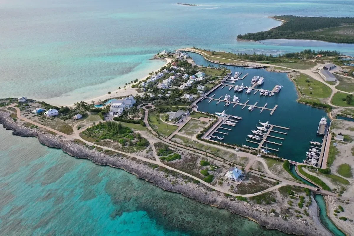 Aerial view of a marina with numerous yachts, houses, and a small beach on an island surrounded by turquoise waters.
