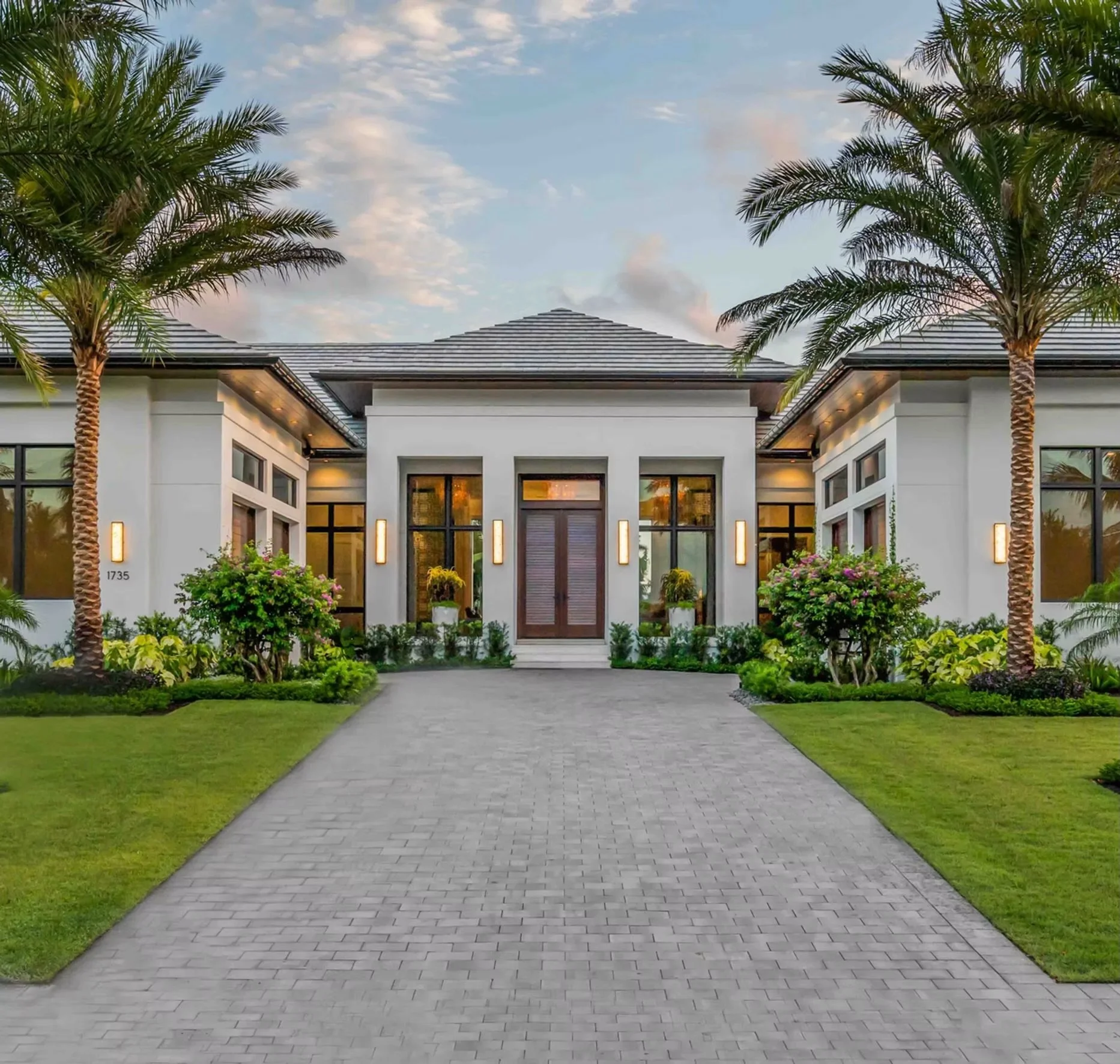 A modern, white, single-story house with large windows, decorative lighting, and landscaped front yard featuring palm trees and green shrubs. A paved driveway leads to the front door.