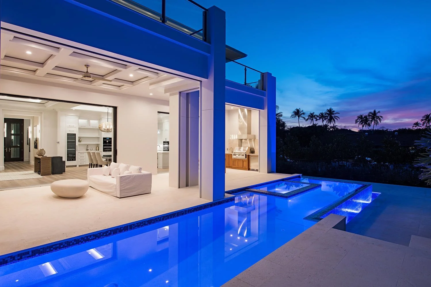 Modern house with open living space featuring a swimming pool and patio area at dusk, with palm trees in the background.