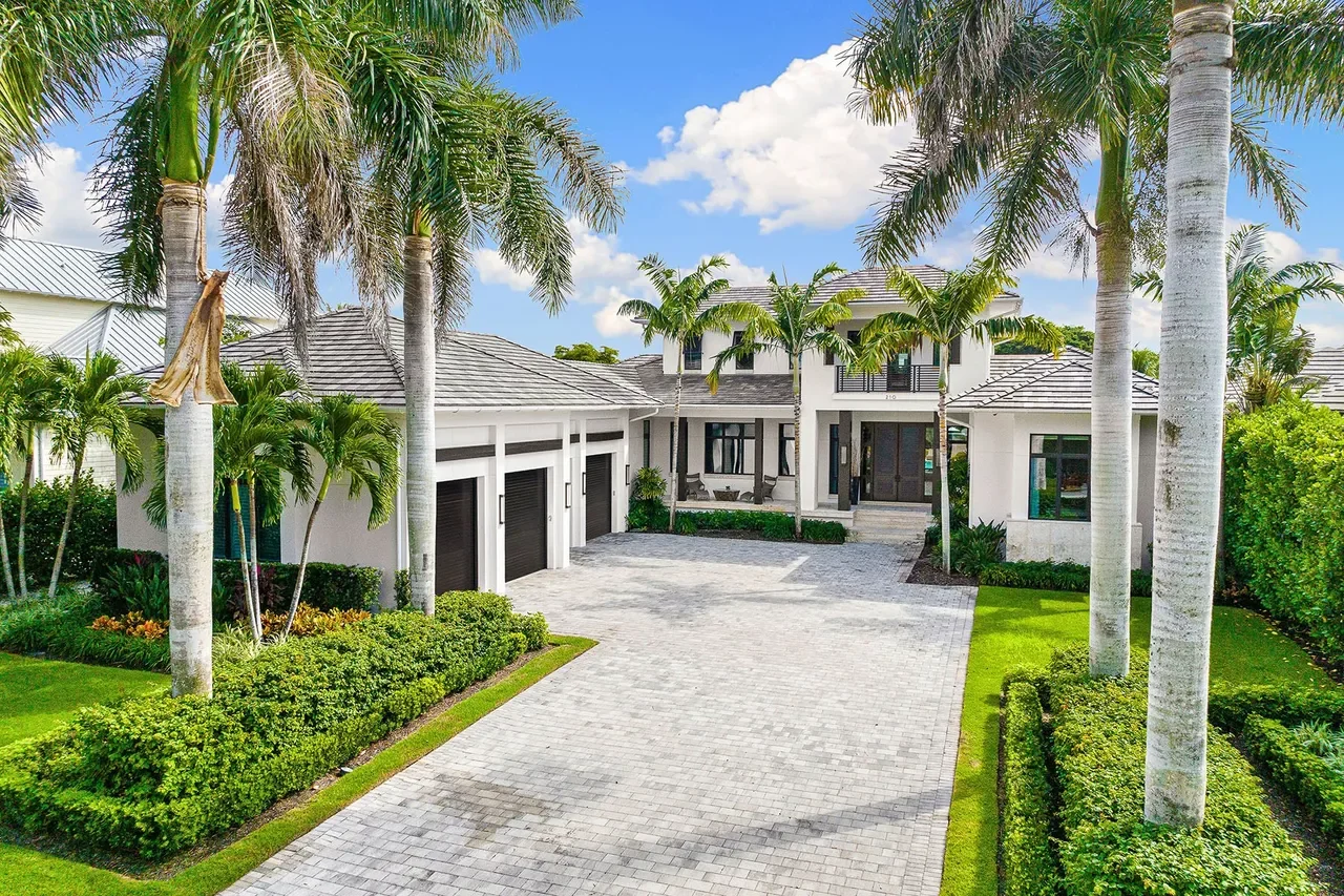 A large modern house with a three-car garage, paved driveway, surrounded by palm trees and well-manicured landscaping, under a partly cloudy sky.