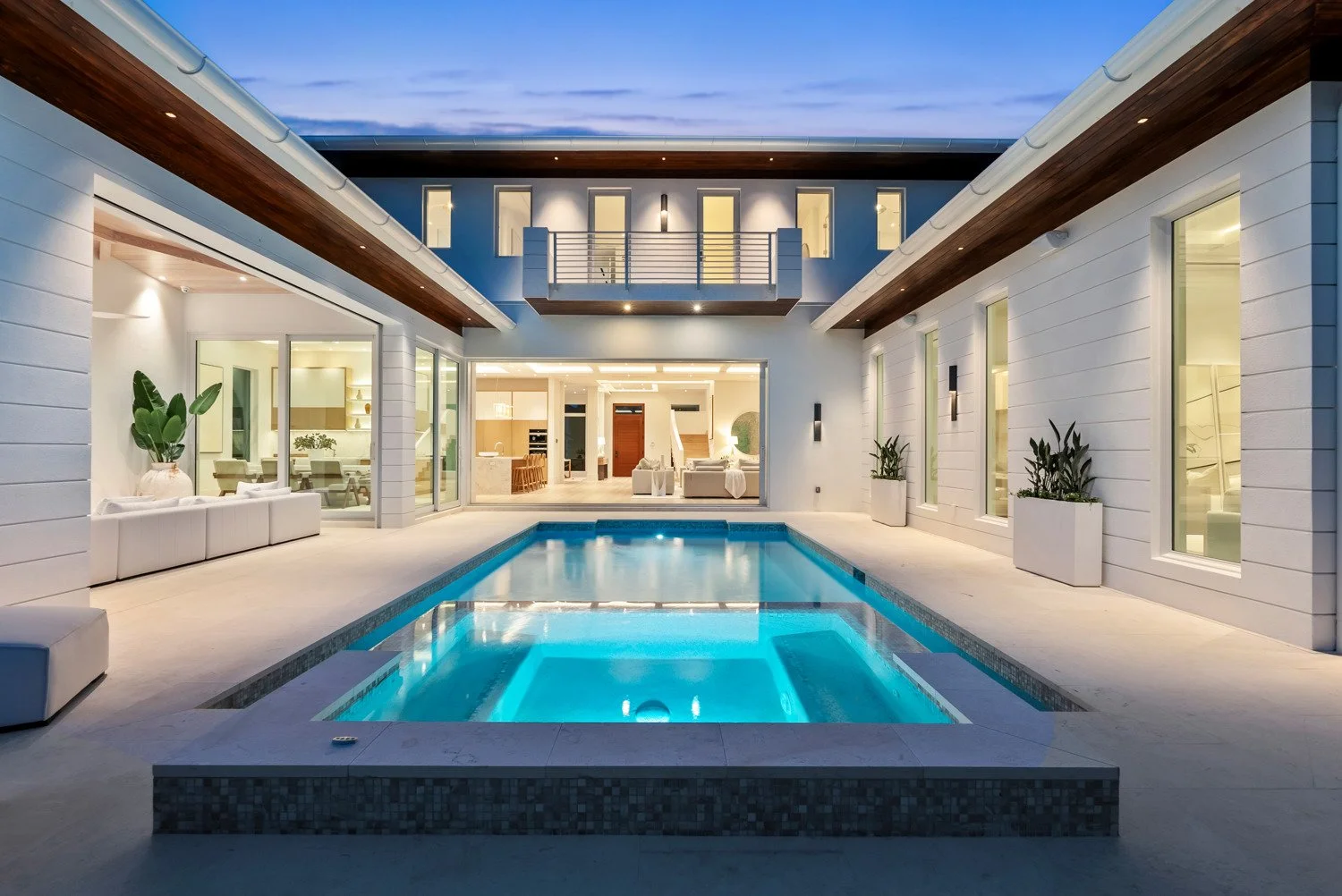Modern backyard with illuminated swimming pool, white patio furniture, potted plants, and a spacious open-plan house interior visible through large glass doors, during dusk.