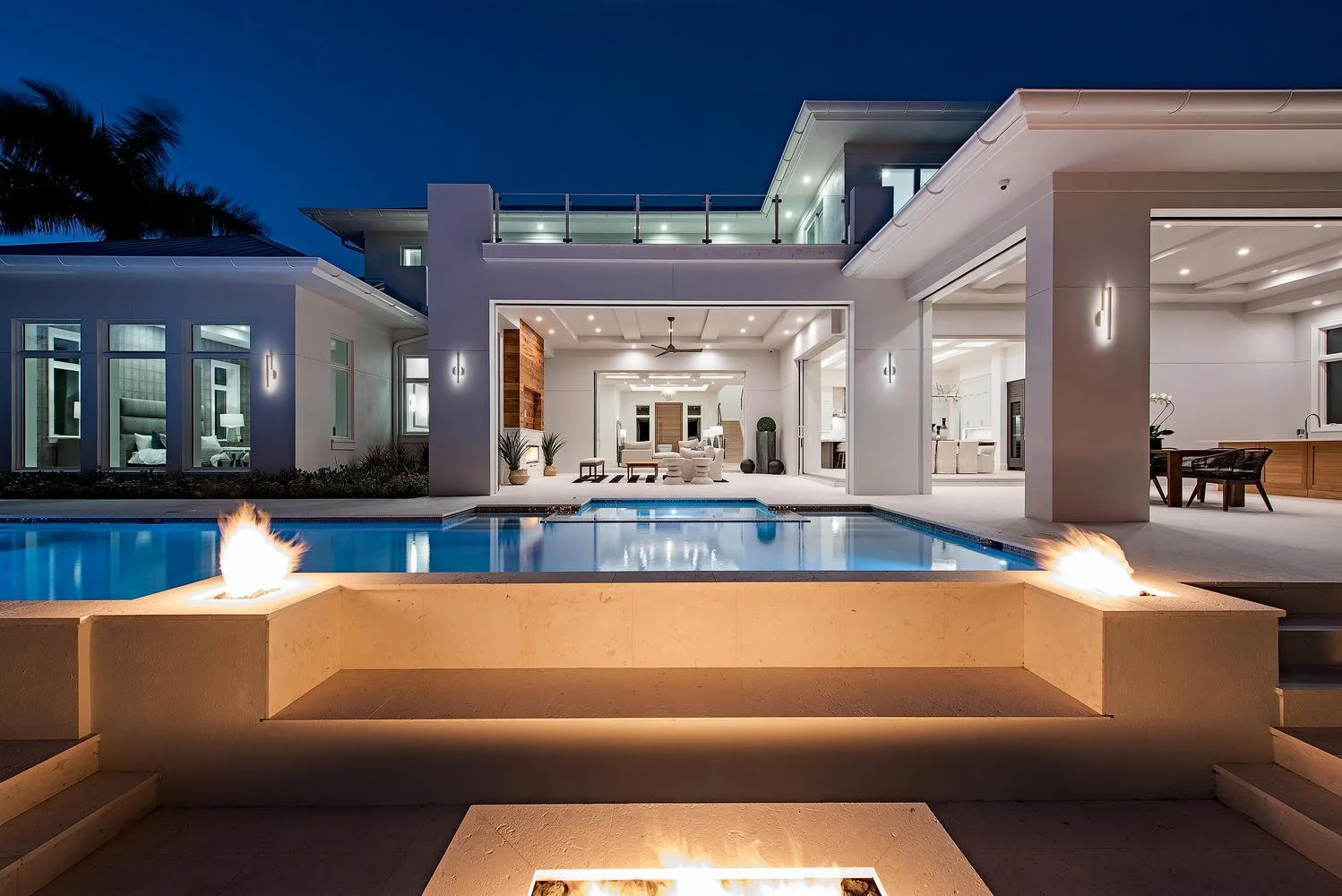 Modern house at night with outdoor pool, fire features, and well-lit interior visible through open spaces.