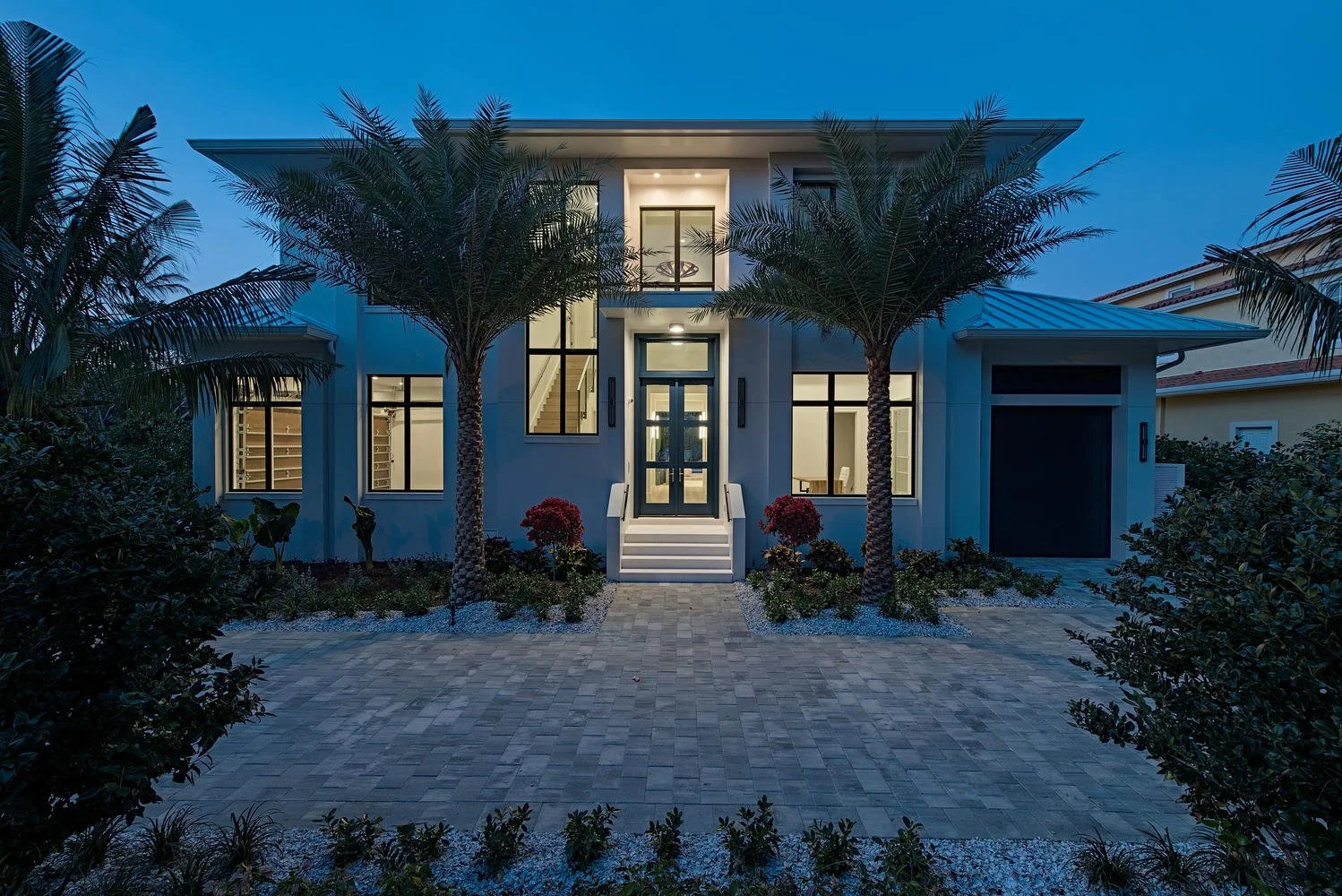 Modern two-story house with large windows, a front porch with stairs, manicured bushes, palm trees, and early evening lighting.