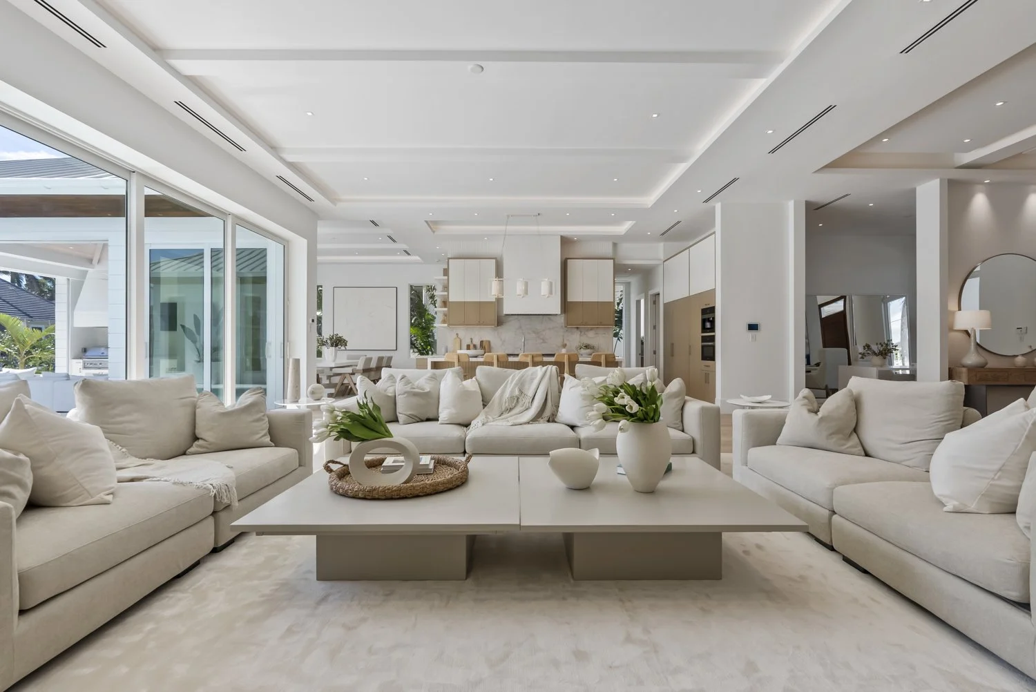 Bright, open-concept living room with white sofas, a large white coffee table, and decorative vases with flowers, overlooking a modern kitchen with white and wood cabinetry.