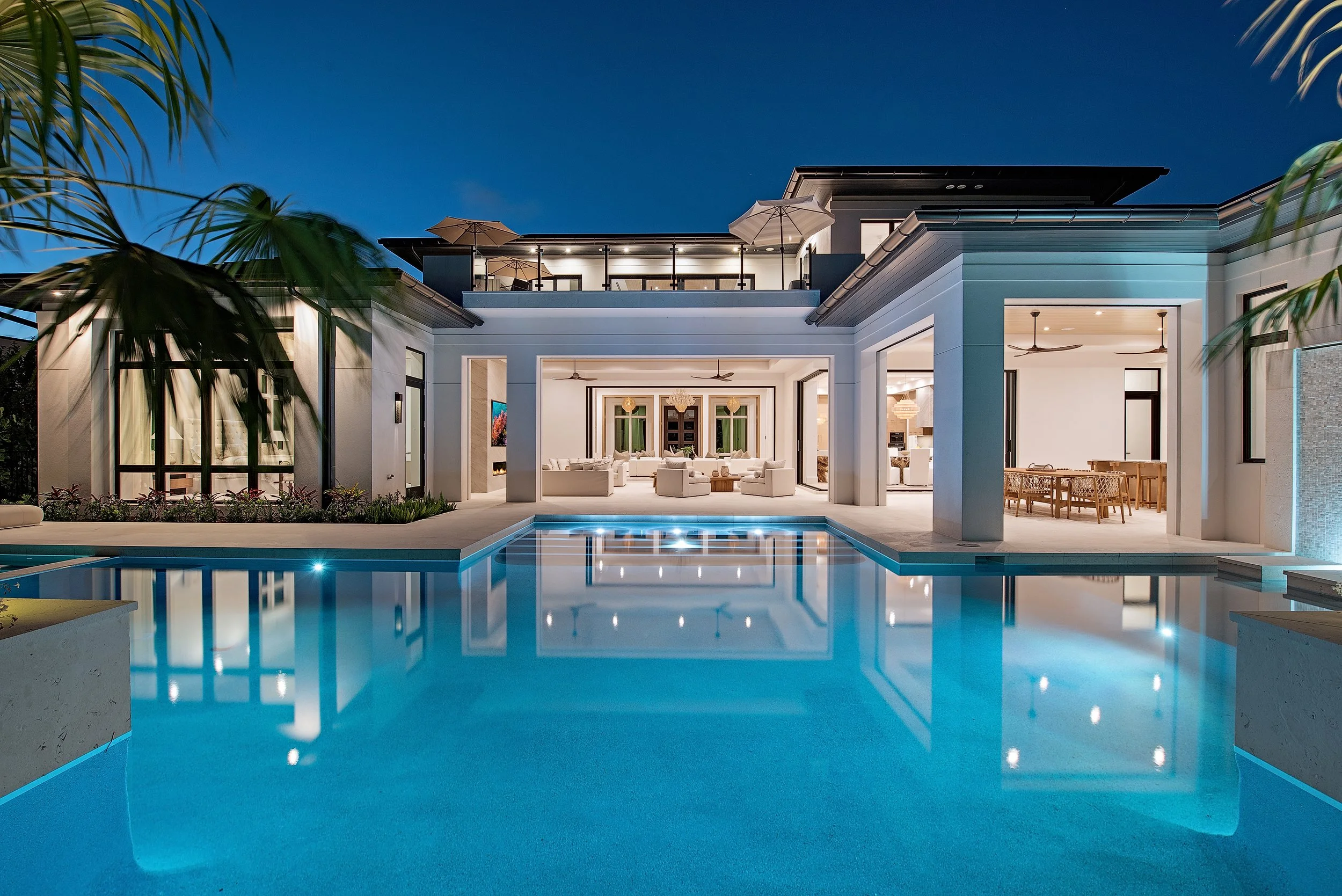 Luxury modern house with illuminated swimming pool at night, open living space, patio with umbrellas, and tropical plants.