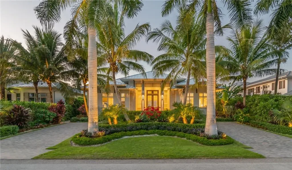 A tropical mansion with lush landscaping, palm trees, and illuminated windows, featuring a stone pathway leading to the entrance.