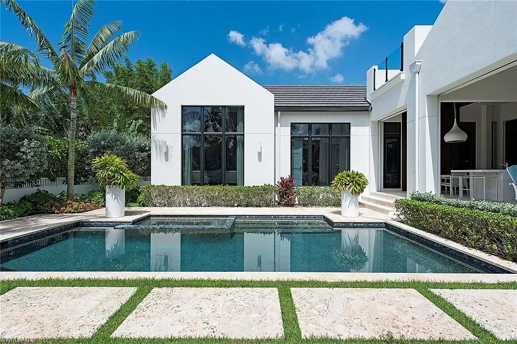 Modern house with a rectangular swimming pool in a landscaped backyard, featuring potted plants, palm trees, and outdoor seating area under a covered patio.