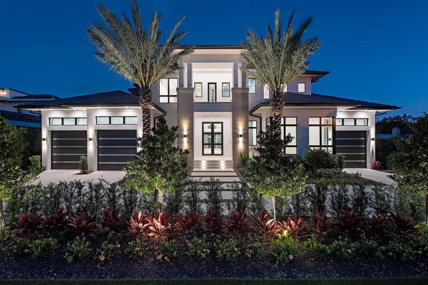 Modern two-story house at night with palm trees and landscaped front yard, exterior lighting, large windows, and garage doors.
