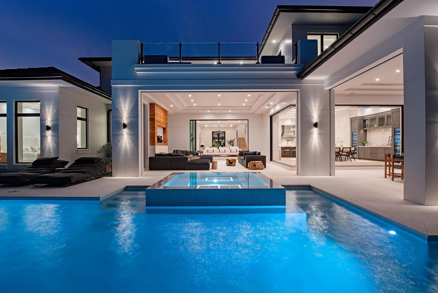 Modern two-story house with a backyard pool and hot tub, illuminated at night, with outdoor lounging areas and a view into the living room and kitchen.