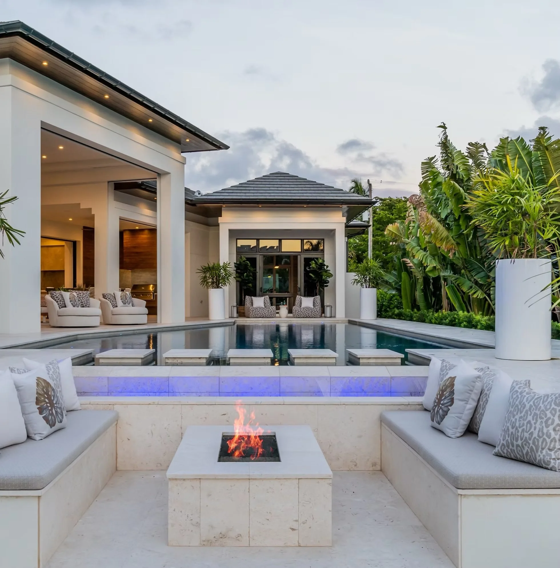 Luxury modern backyard with a pool, outdoor seating with cushions, and a fire pit, surrounded by lush tropical plants.