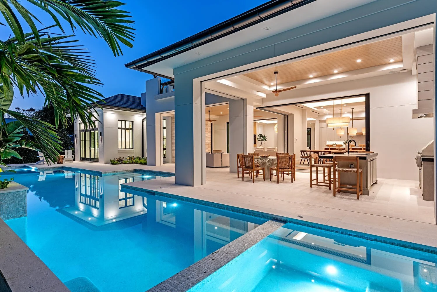 Modern house with a swimming pool, illuminated outdoor patio with dining area, ceiling fans, and indoor lighting visible through large open sliding doors at dusk.