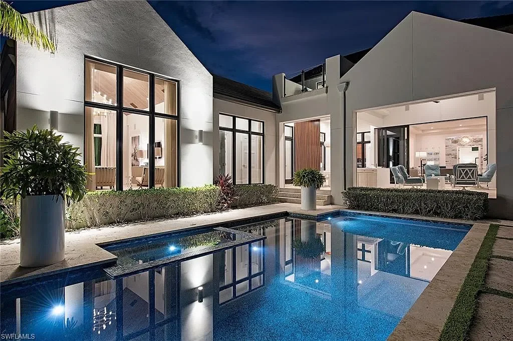 Modern house with large windows at night, overlooking a lit swimming pool with clear water, surrounded by landscaped plants and outdoor furniture visible through open windows.
