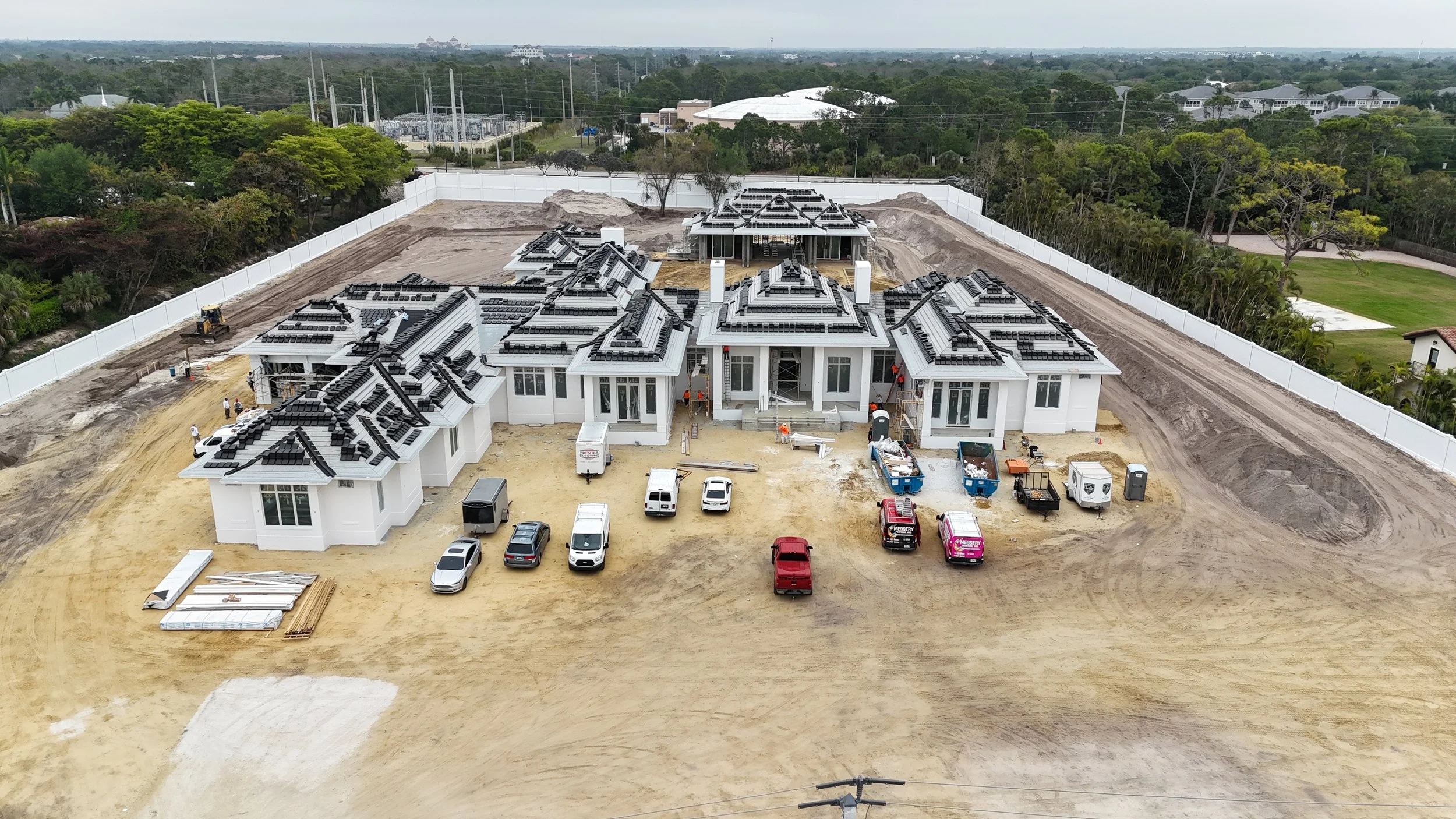 A large house under construction with a white exterior and a complex roof design. The house is surrounded by a dirt lot with construction vehicles and equipment, and a white fence encloses the property. There are multiple cars parked in front of the house, and workers are visible on site.