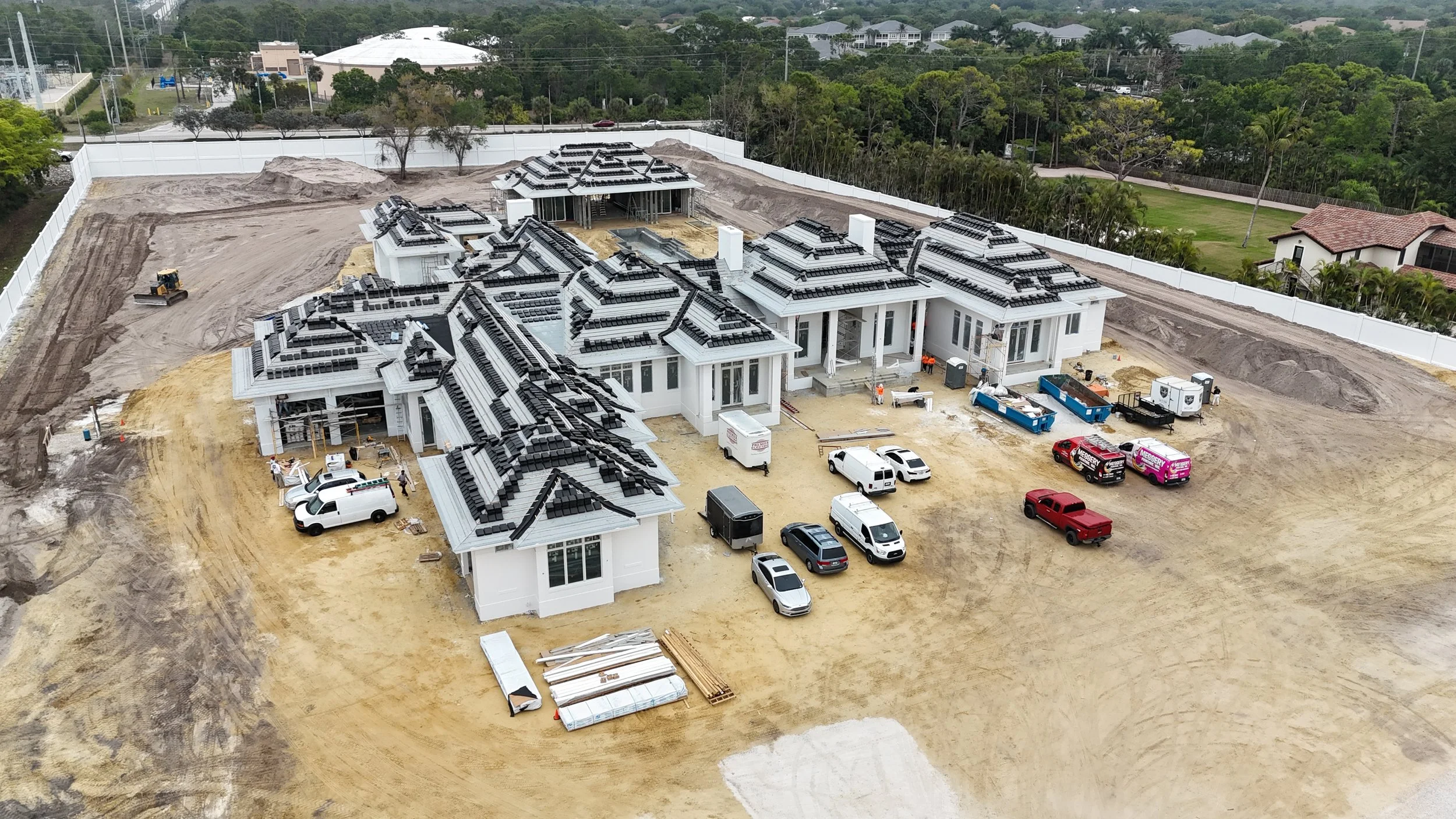 A large residential building under construction with multiple sections, solar panels on the roofs, and surrounding construction site with vehicles, equipment, and materials.