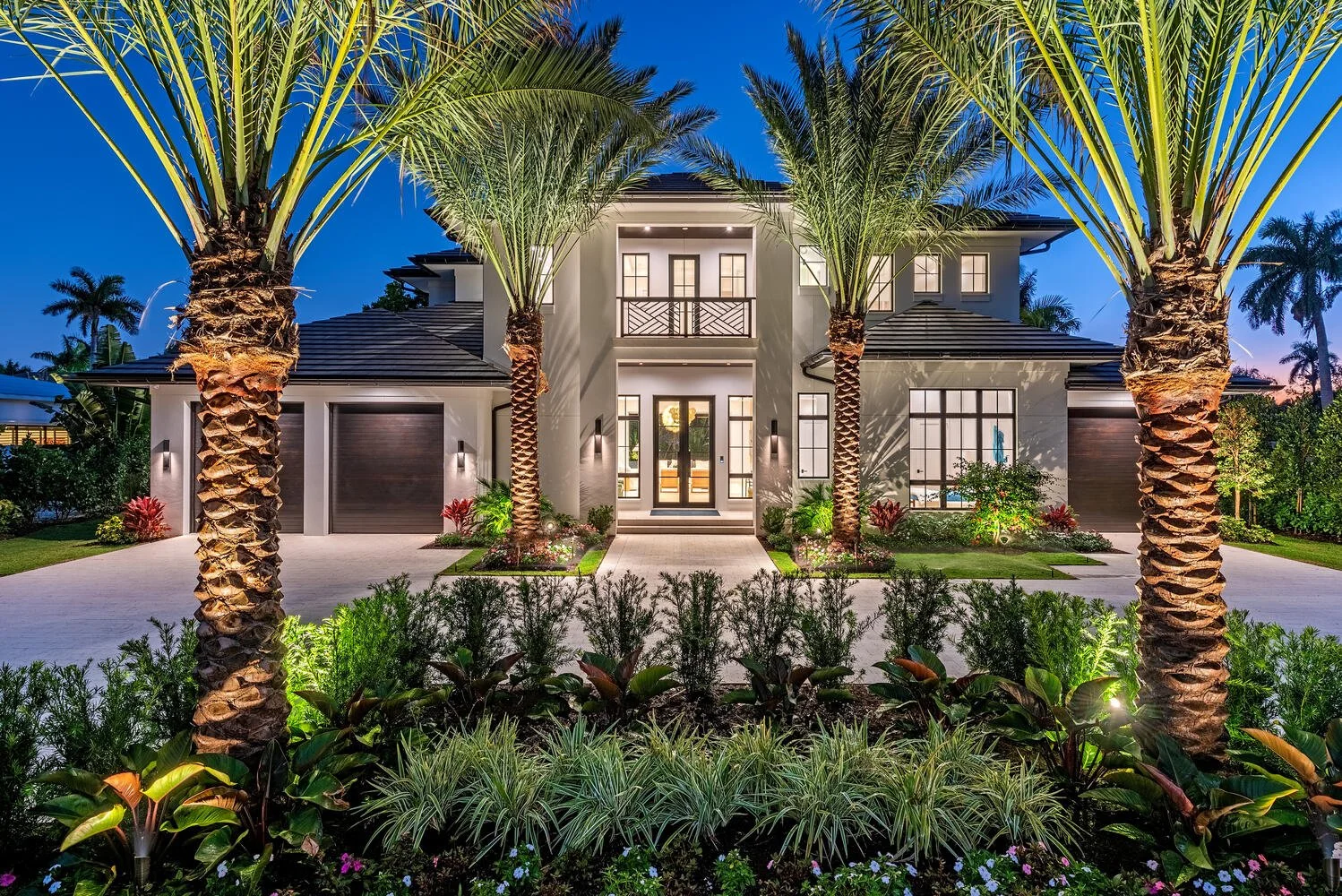 A modern two-story house illuminated at night, with a well-maintained front yard featuring palm trees, shrubs, and flowers, a paved driveway, large windows, and a front porch.