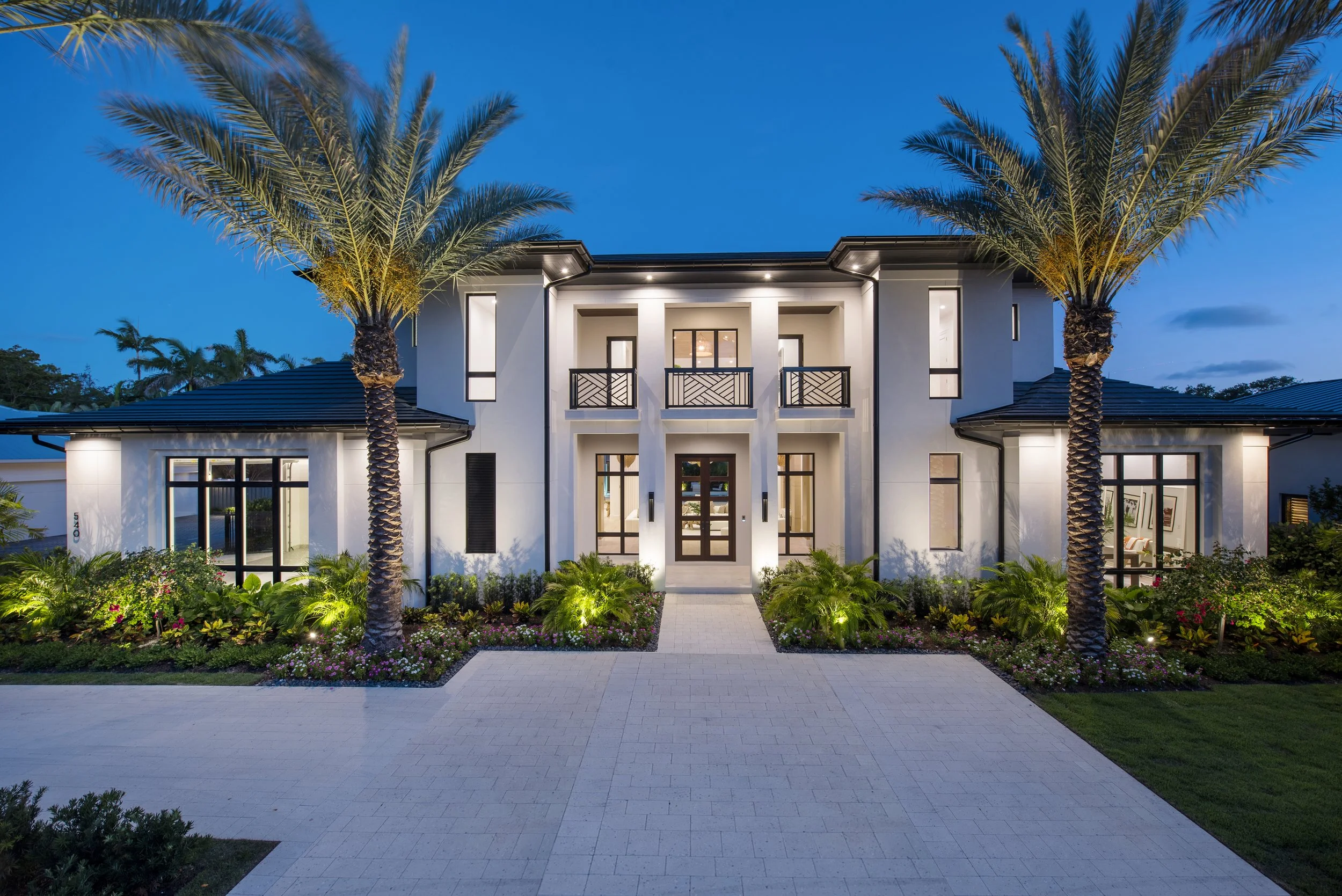 Modern two-story house with white exterior walls, dark roof, and symmetrical design. Front entrance features a wooden door, surrounded by large glass windows. Two tall palm trees are on either side of the walkway, with well-maintained landscaping and garden beds. The house is lit up at dusk.