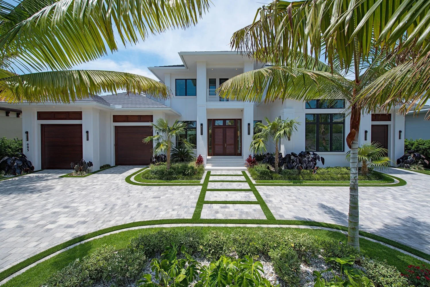 Front view of a modern white two-story house with large windows, a paved driveway, and lush tropical landscaping, including palm trees and green shrubs.