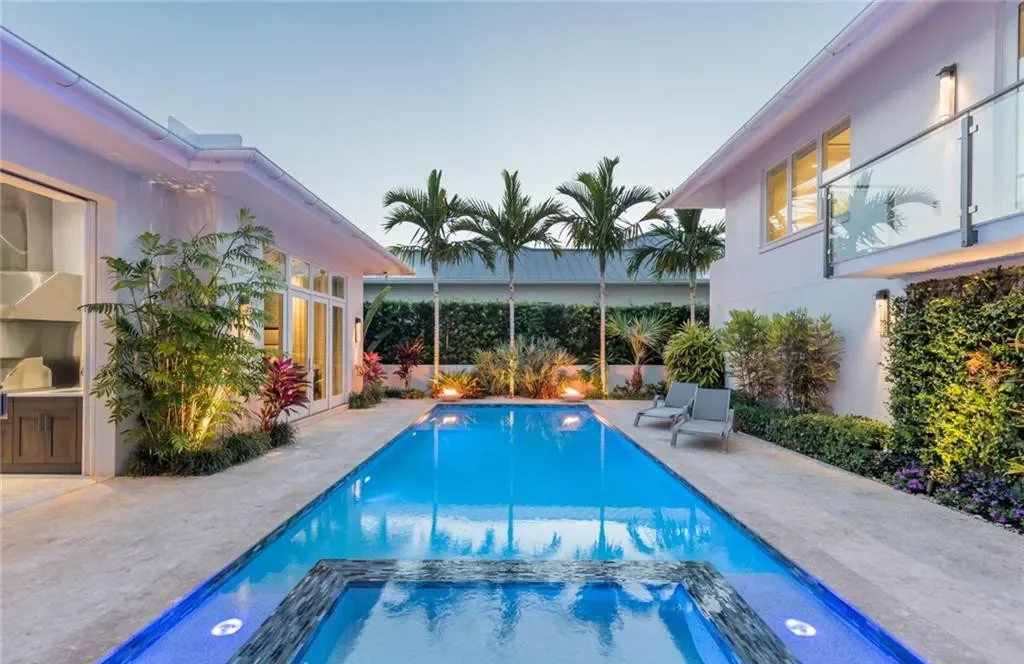 A backyard with a rectangular swimming pool, surrounded by outdoor lounge chairs, lush plants, and palm trees, with a modern white house in the background.