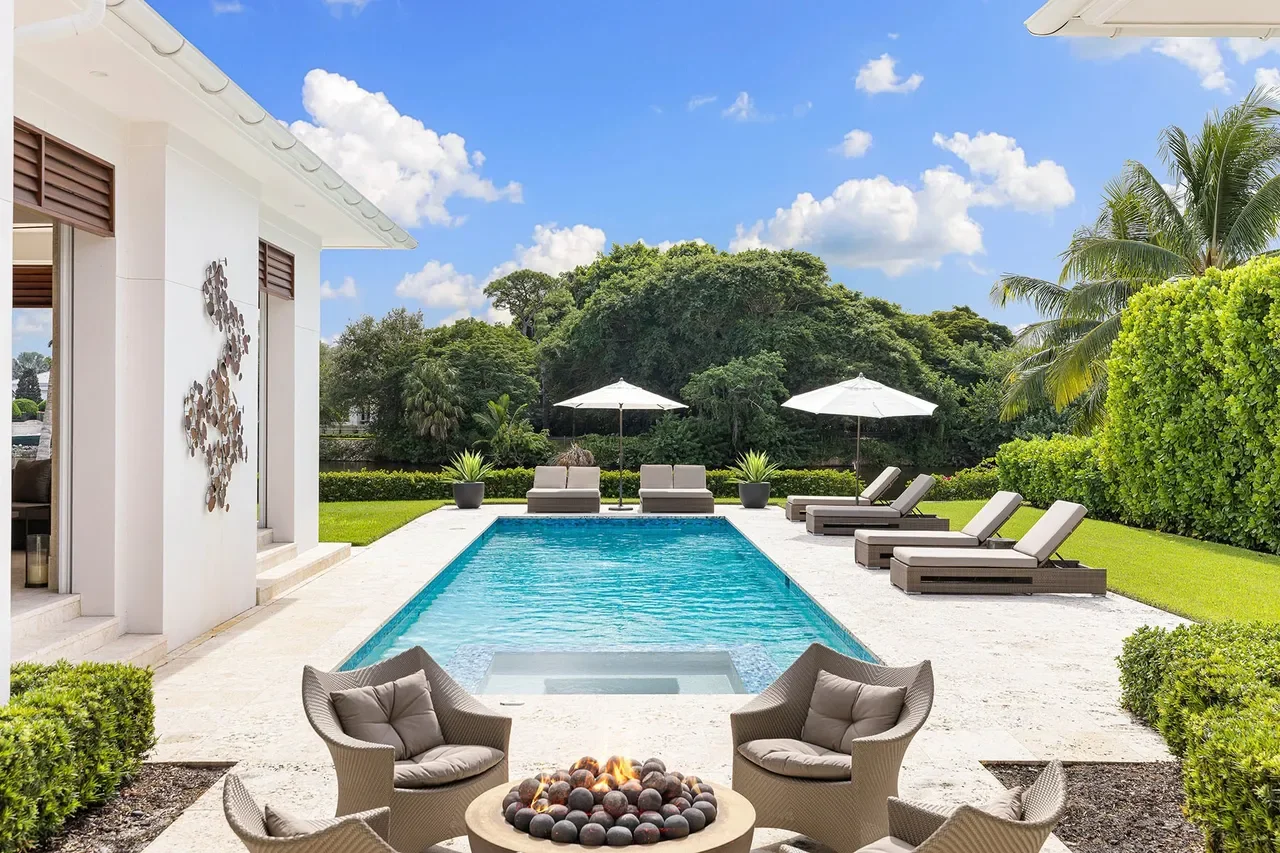 A backyard with a swimming pool, lounge chairs, umbrellas, and seating area, surrounded by greenery and trees under a partly cloudy sky.