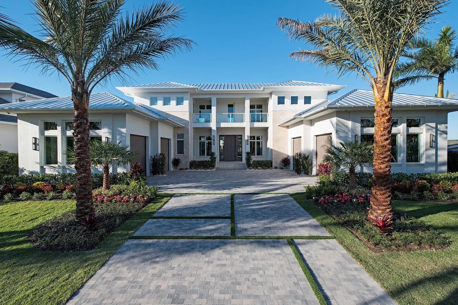 Modern white house with a landscaped front yard, palm trees, colorful flowers, a paved walkway leading to the entrance, and clear blue sky.