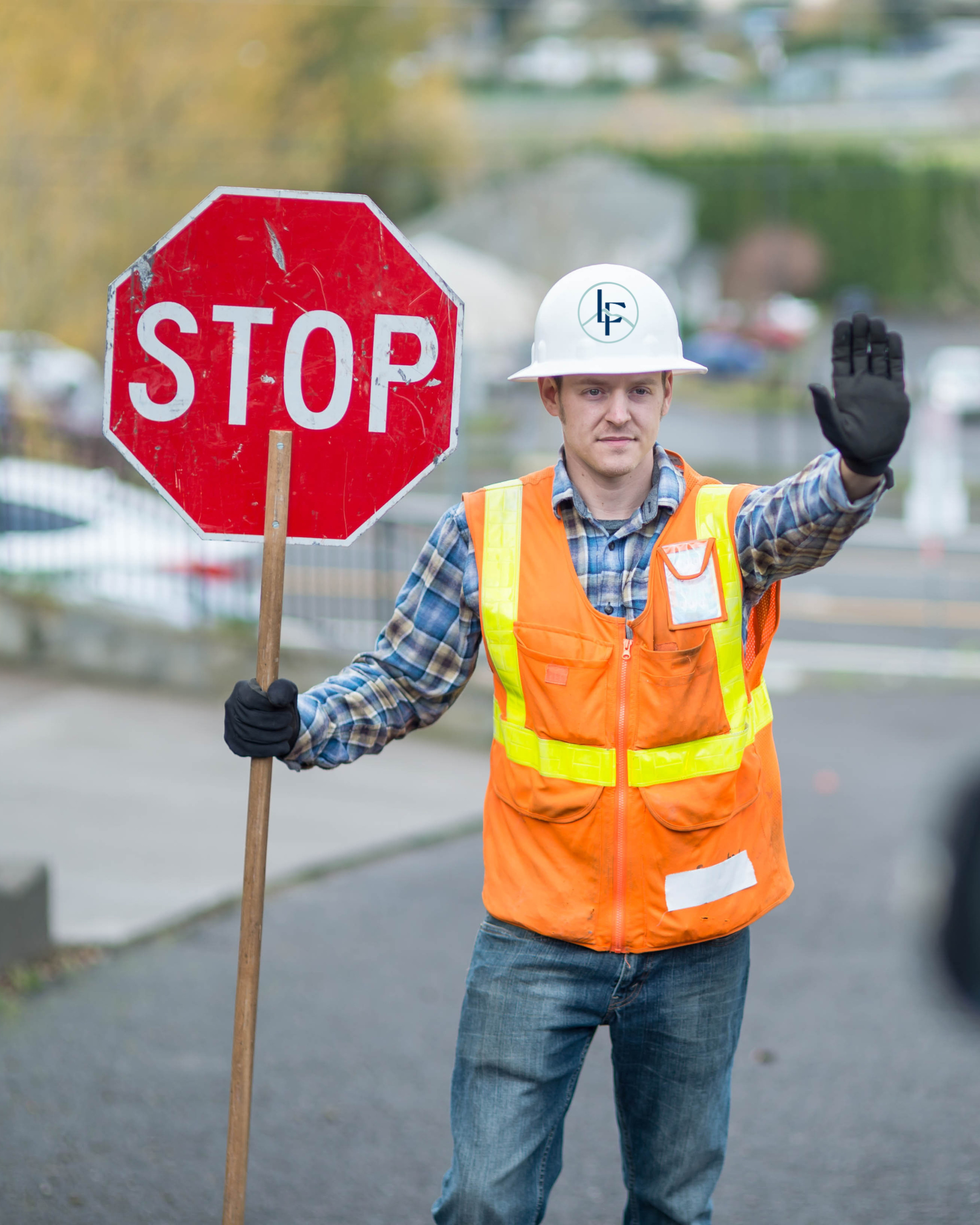 A road construction worker wearing a hard hat and high-visibility vest stands in the street holding a red octagonal sign that reads “STOP” while raising one hand to signal traffic to stop.