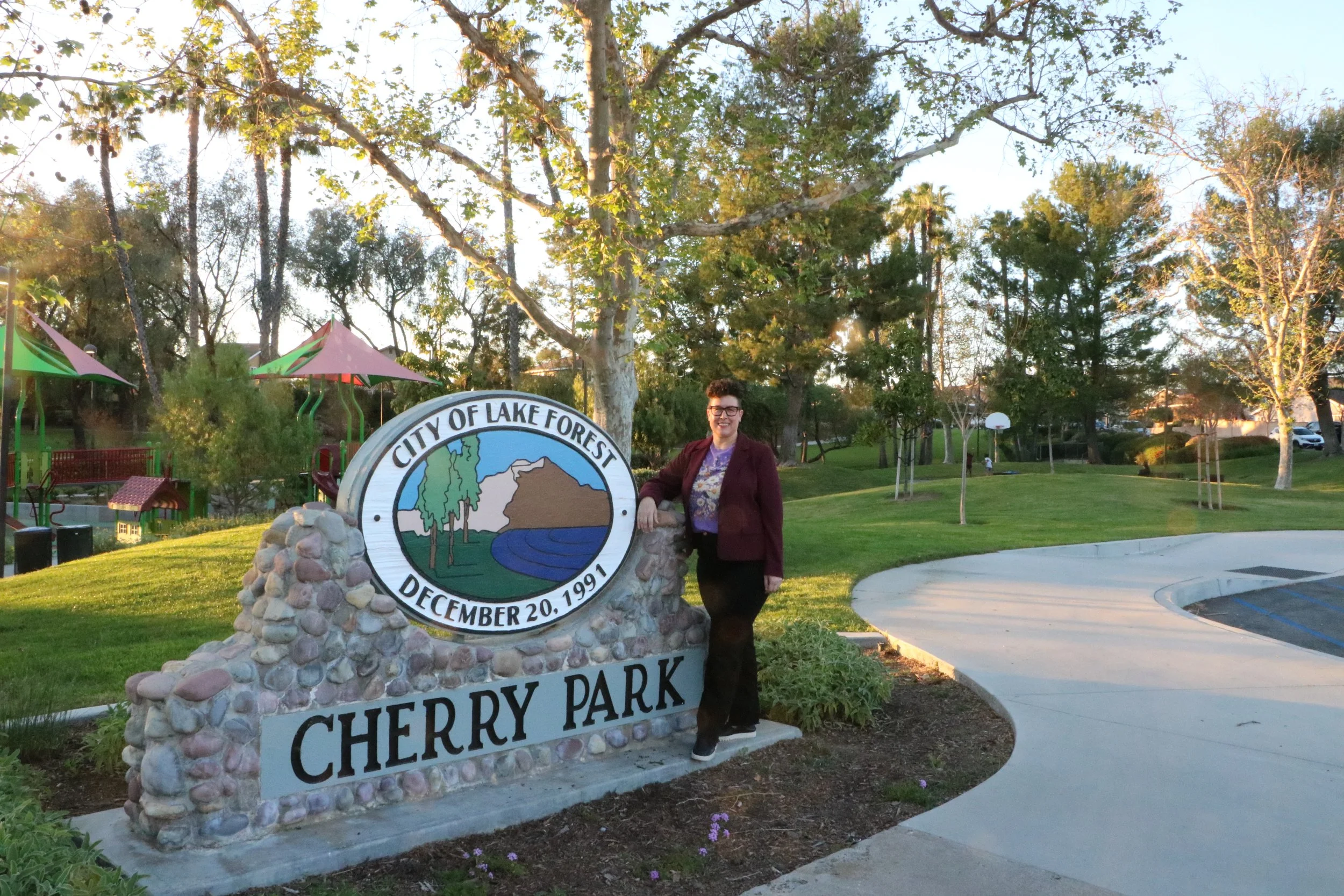 A person stands beside a stone sign that reads “City of Lake Forest December 20, 1991 Cherry Park” in a grassy park with trees, a playground, a basketball court, and a curved walking path in the background.