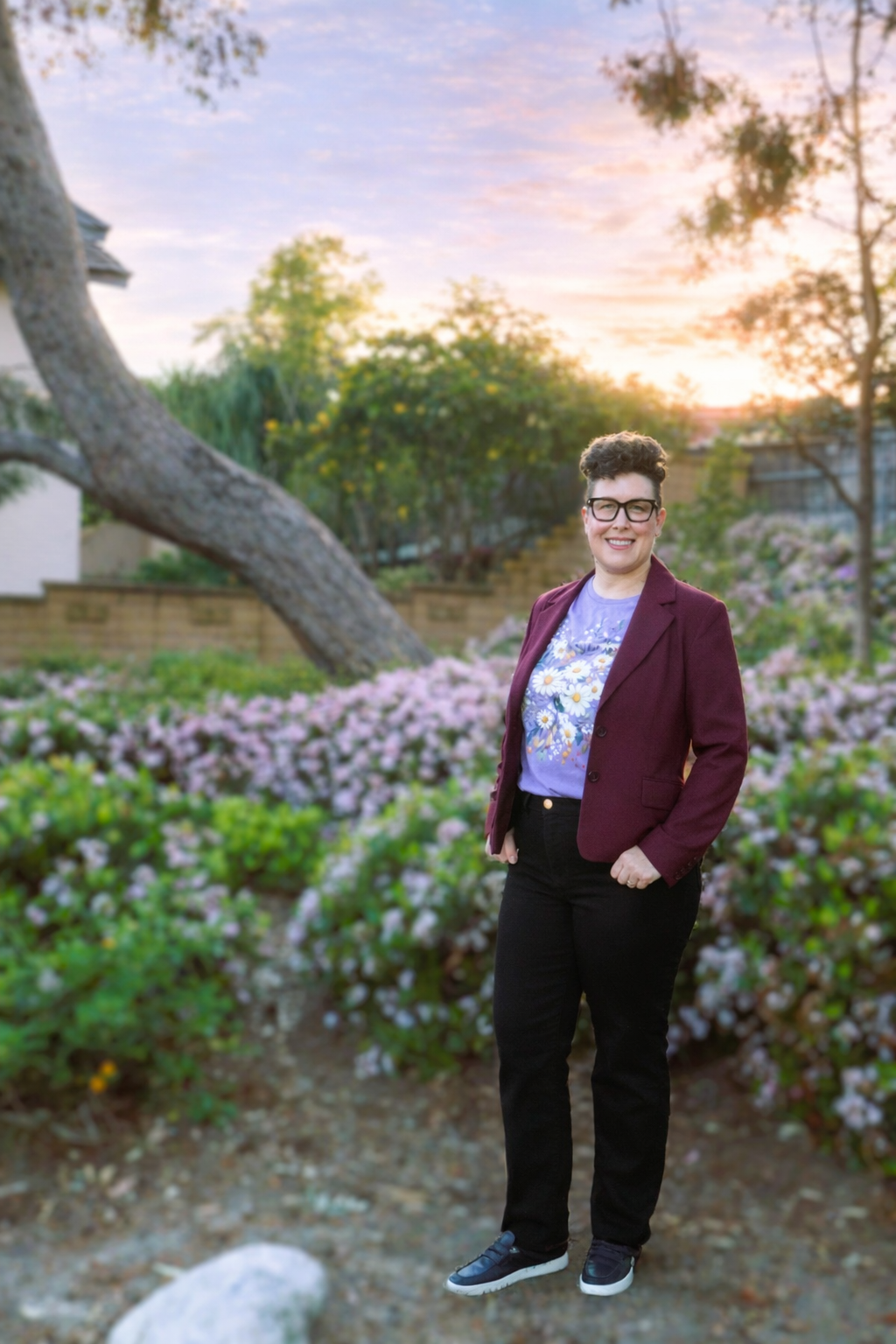 Amy stands smiling in a garden at sunset, wearing glasses, a blazer, a graphic T-shirt, and dark pants. She poses near flowering shrubs and a curved tree, with a residential fence and soft sky in the background.