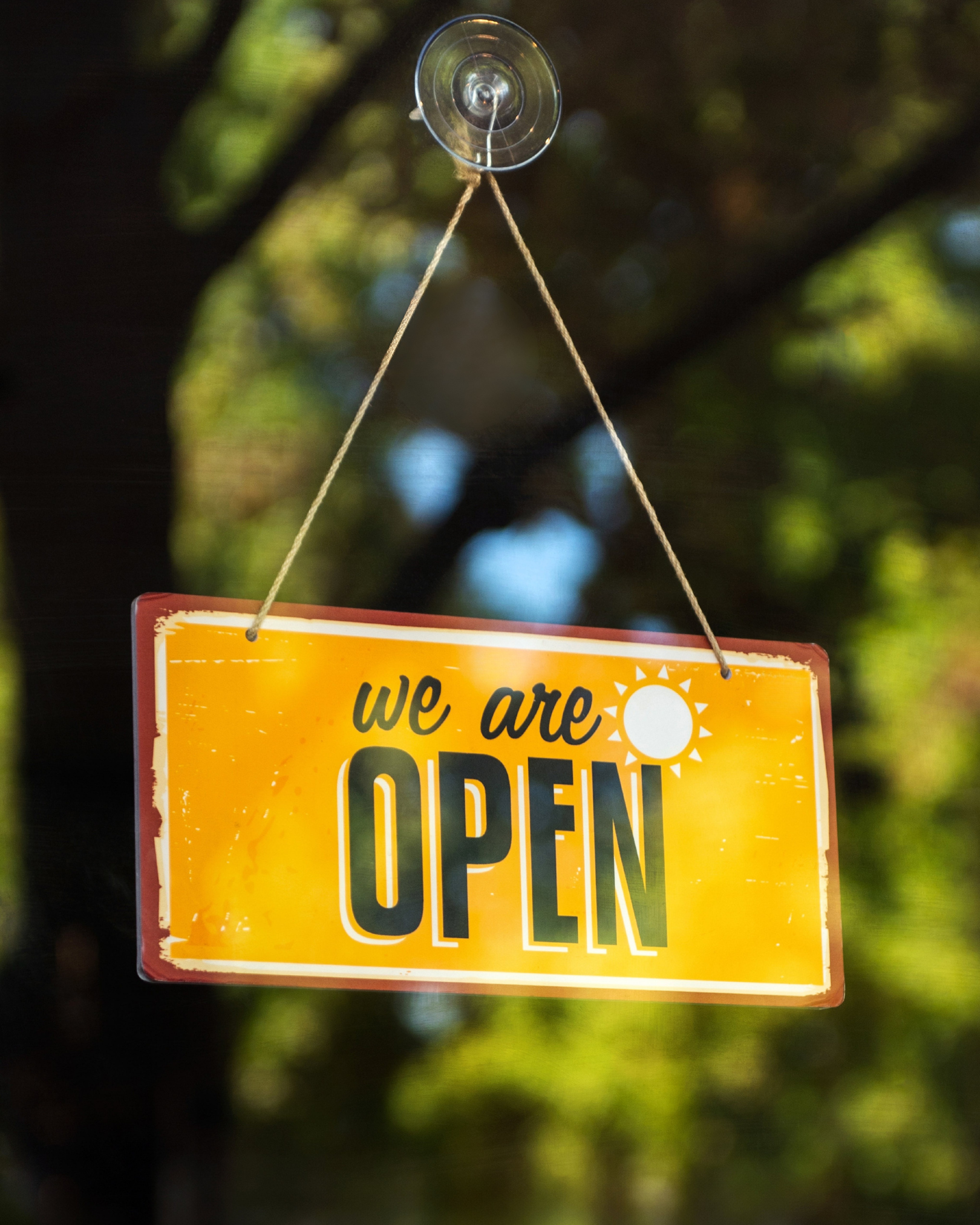 A yellow rectangular sign hangs by two ropes from a suction cup on glass. The sign reads “We are OPEN” in large letters with a small sun icon, with blurred green trees in the background.
