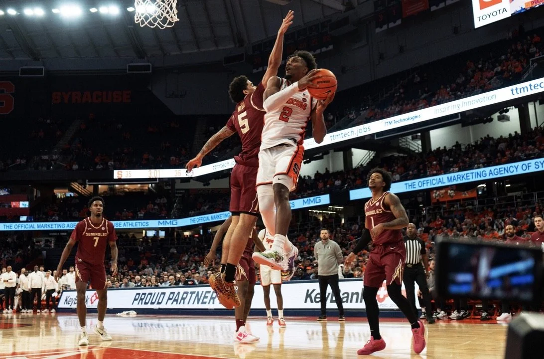 Two basketball players, one from Syracuse in a white jersey and one from Florida State in a maroon jersey, jump to contest a shot at the basket during a game. The Syracuse player is holding the ball while the Florida State player attempts to block.
